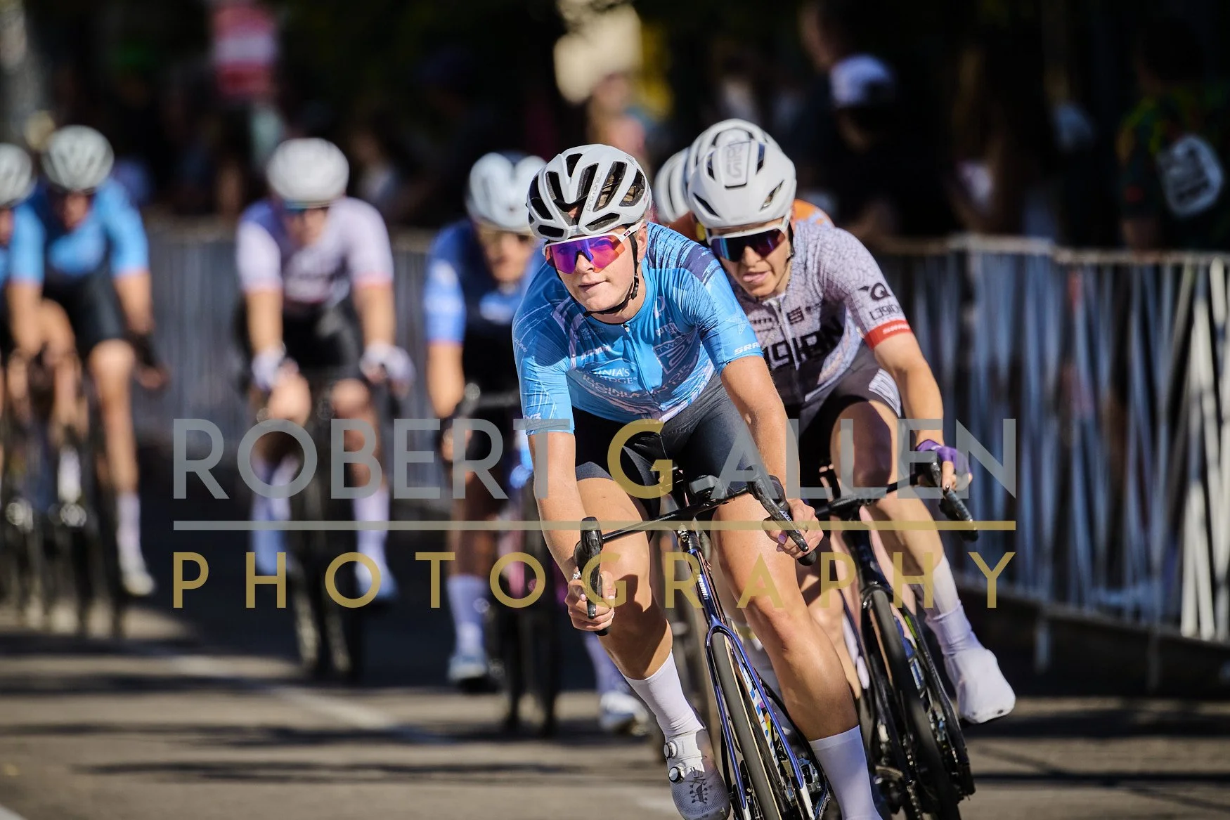 Cyclists participating in a race, wearing helmets and sports uniforms, riding on a road with spectators behind a barrier in the background.