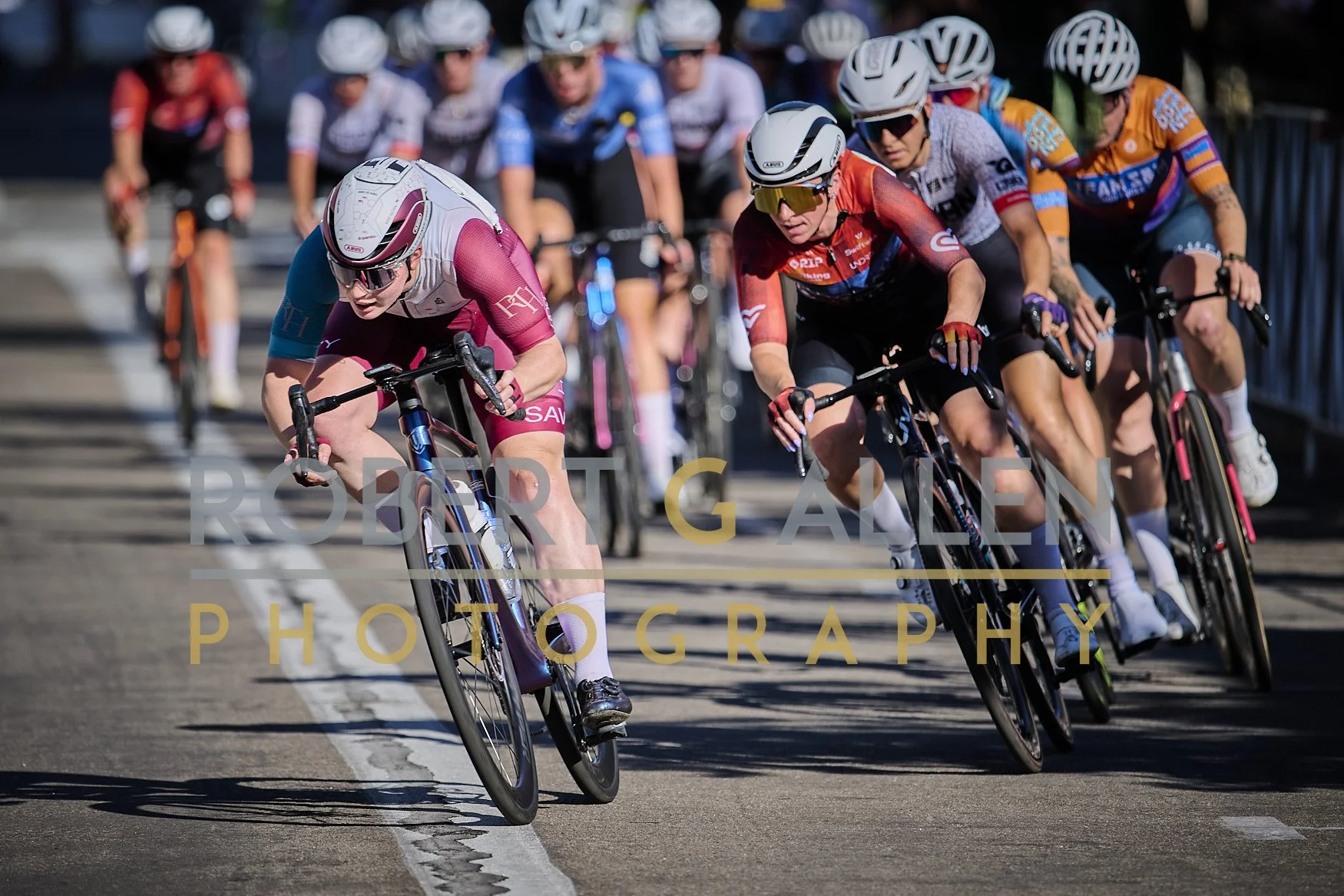 A group of cyclists racing on a road, with some leaning into a turn, wearing colorful jerseys, helmets, and sunglasses.