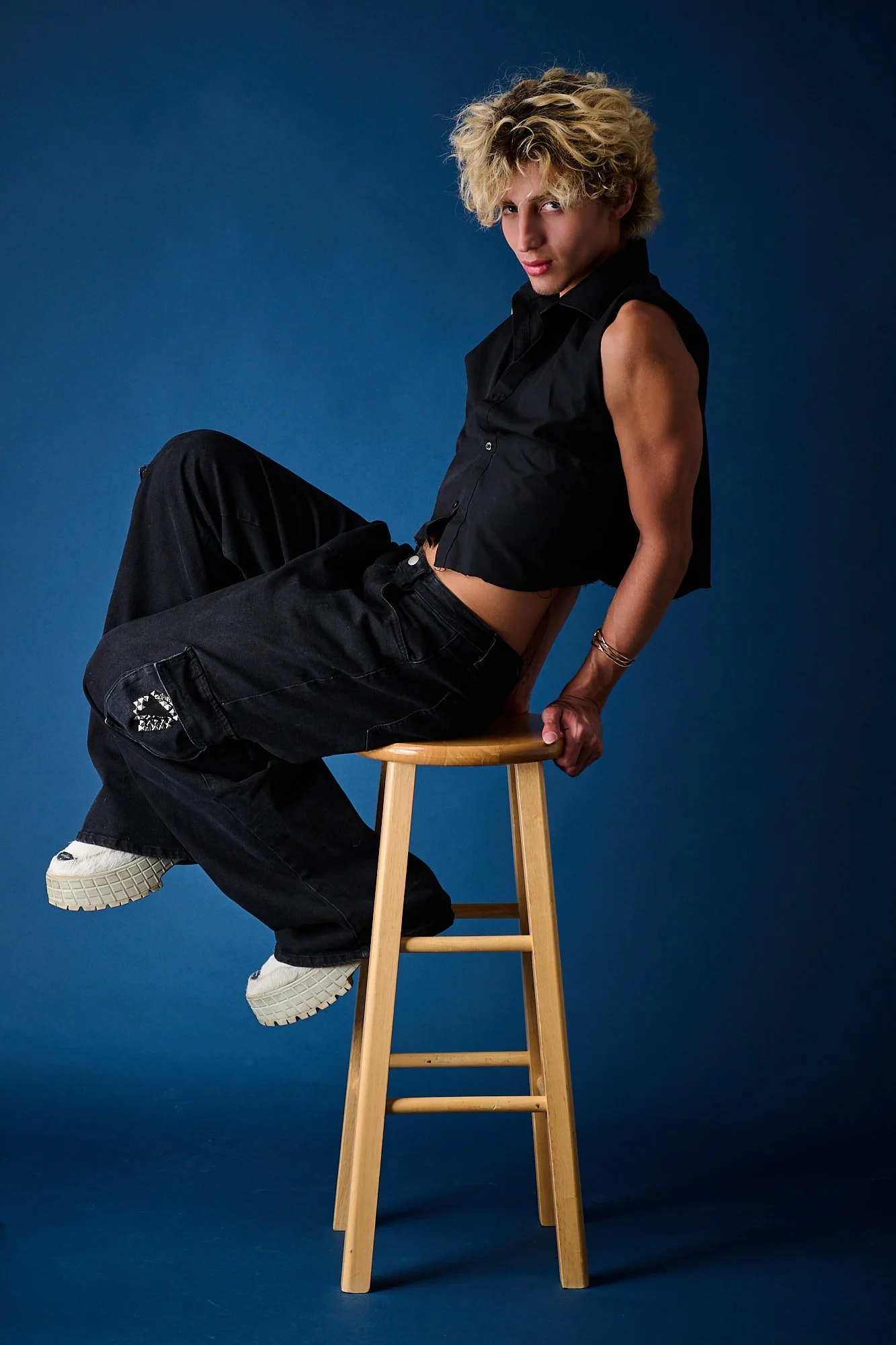 A young man with curly blond hair sitting sideways on a wooden stool against a blue background. He wears a black sleeveless shirt, black pants, and white sneakers, with one hand resting on the stool. He looks directly at the camera with a confident e