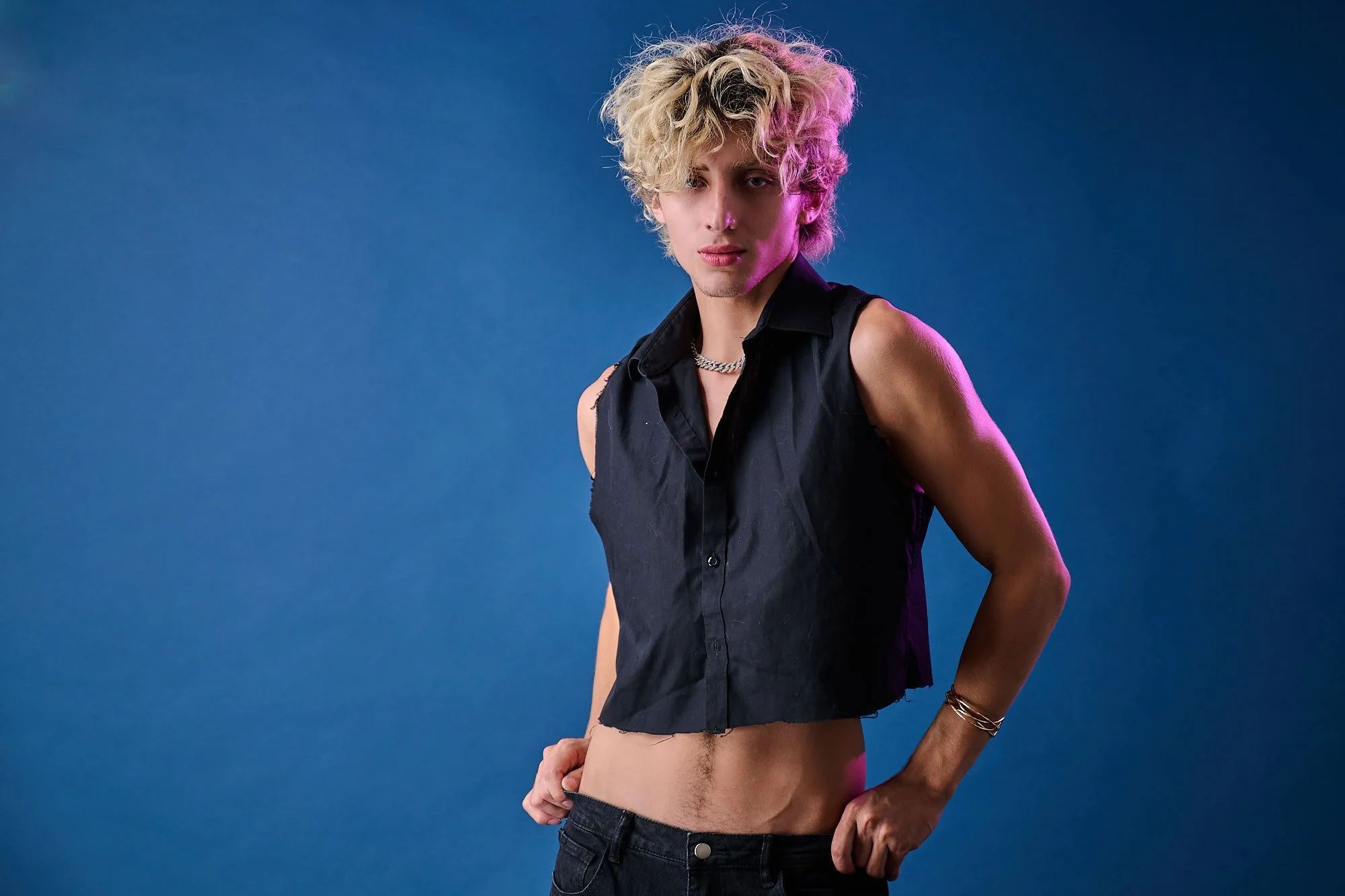 Young man with curly blond hair, wearing a sleeveless black vest, silver chain necklace, and multiple bracelets, topless and with partially unbuttoned jeans, standing against a blue background.