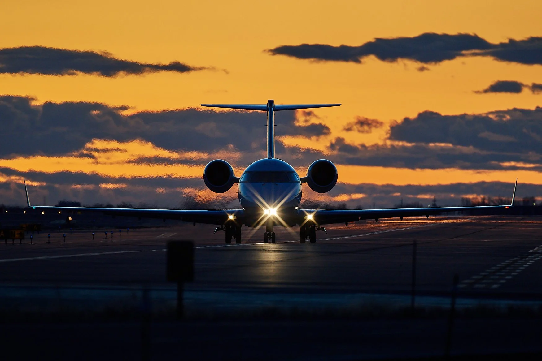 A private jet on an airport runway at sunset with its headlights on, dark clouds in the sky, and the sun setting in the background.