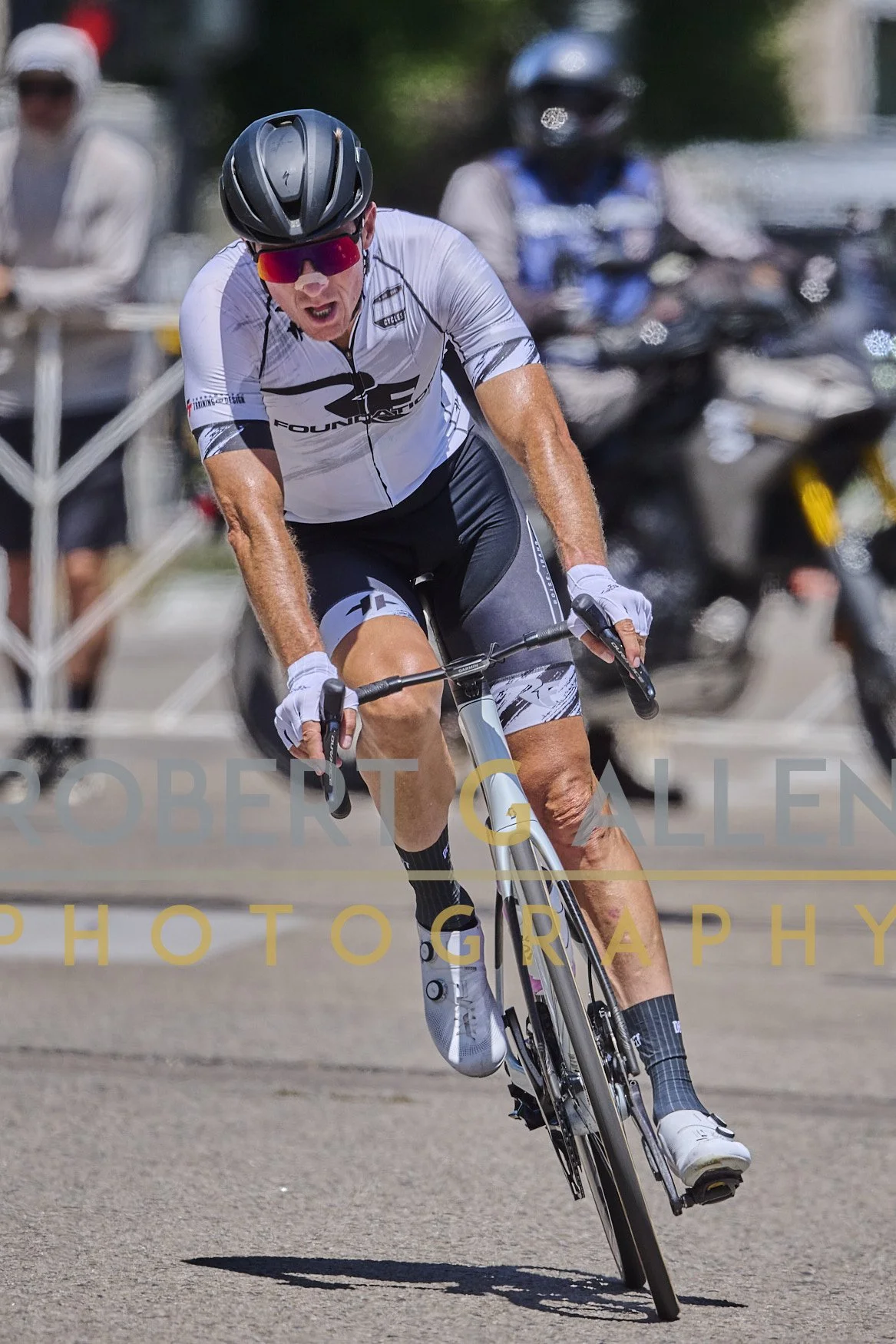 A cyclist wearing a black helmet, red visor, white and black cycling outfit, and sunglasses riding a bike on a paved street. The cyclist appears to be focusing intensely during a race or ride, with a blurred background of onlookers and motorcycles.