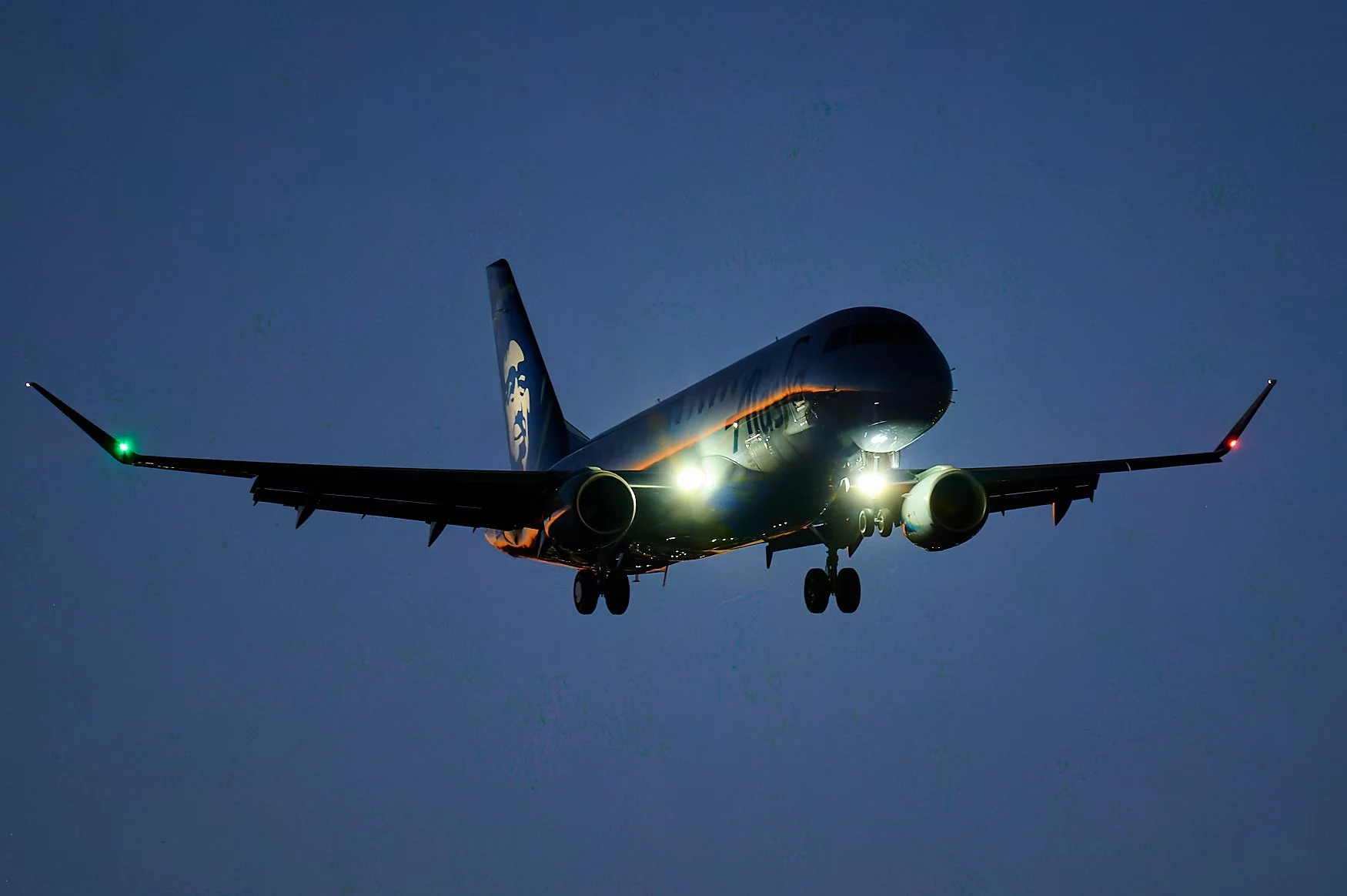 A commercial airplane flying at night with its landing lights on and green and red navigation lights illuminated.