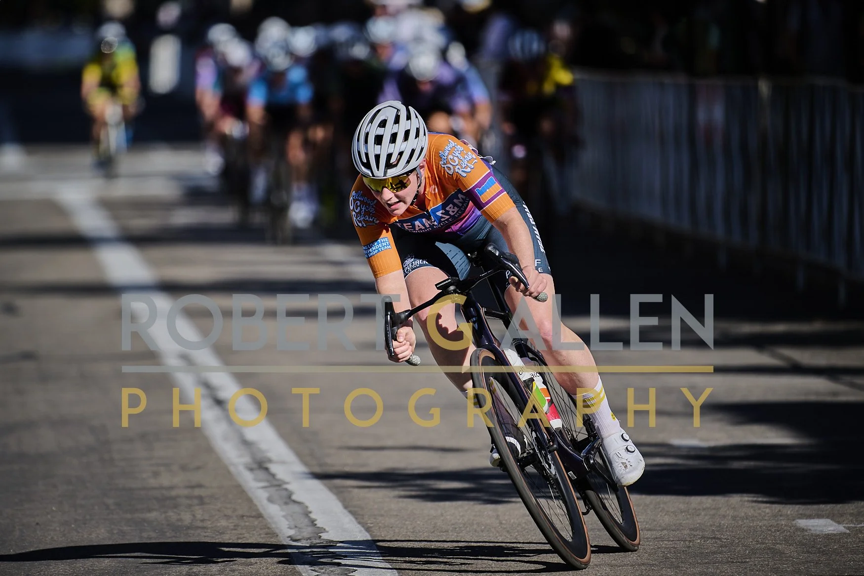 Cyclist racing on a paved road, leaning sharply into a turn, wearing an orange jersey, helmet, and sunglasses, with a group of riders in the background.