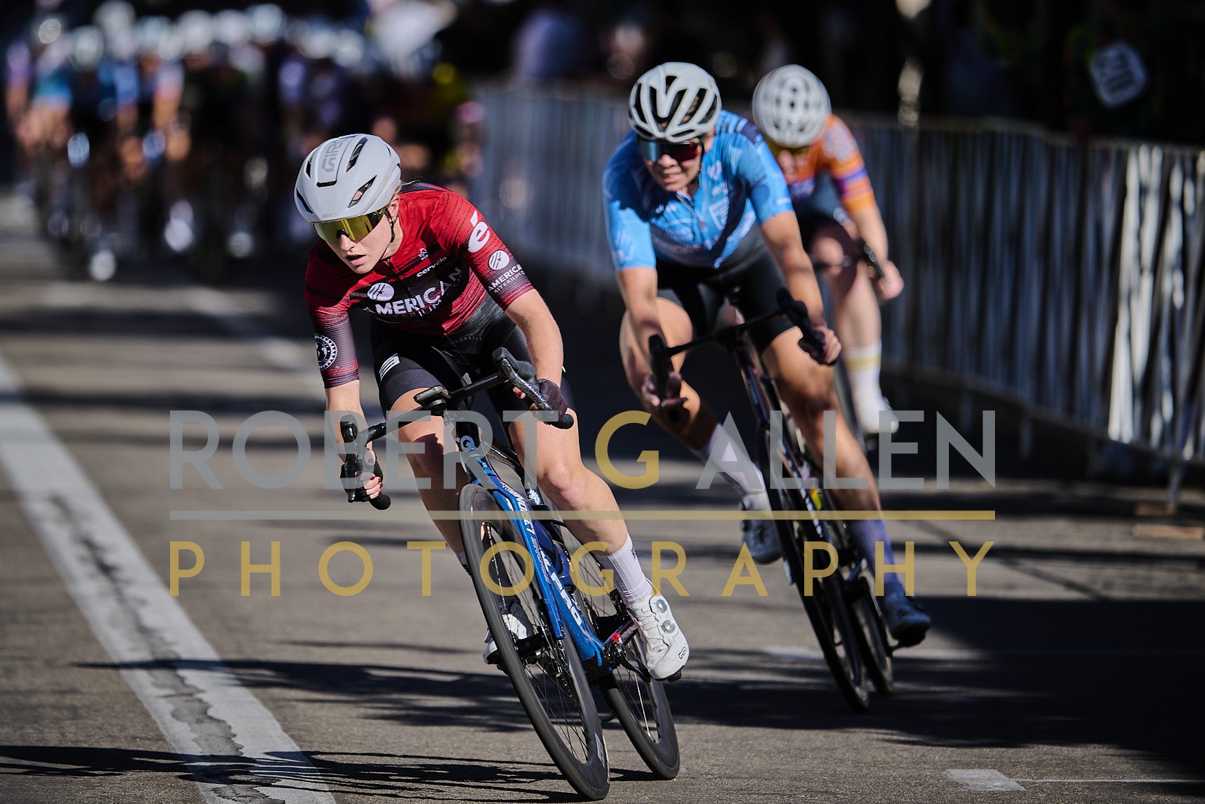 Cyclists competing in a race on a city street, wearing helmets and colorful jerseys, with a blurred crowd in the background.