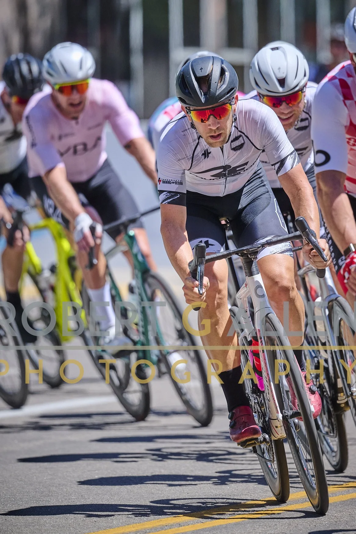Group of cyclists racing on a road during daytime, wearing helmets and sunglasses, with one rider in the front leaning forward and pedaling intensely.