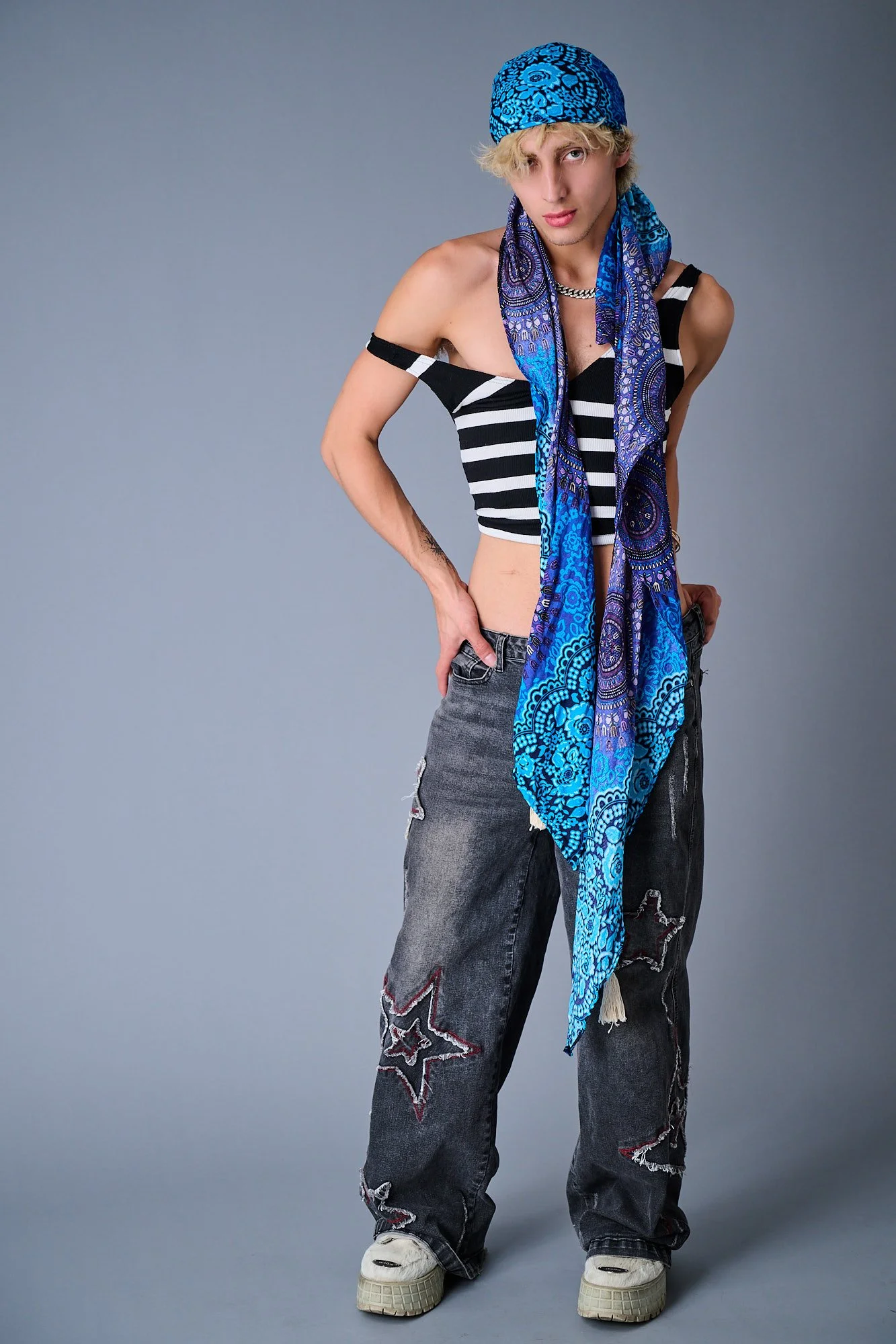 A person with blonde hair wearing a blue patterned bandana, a black and white striped crop top, oversized baggy jeans with star embroidery, white sneakers, and a blue paisley scarf around their neck, standing against a gray background.