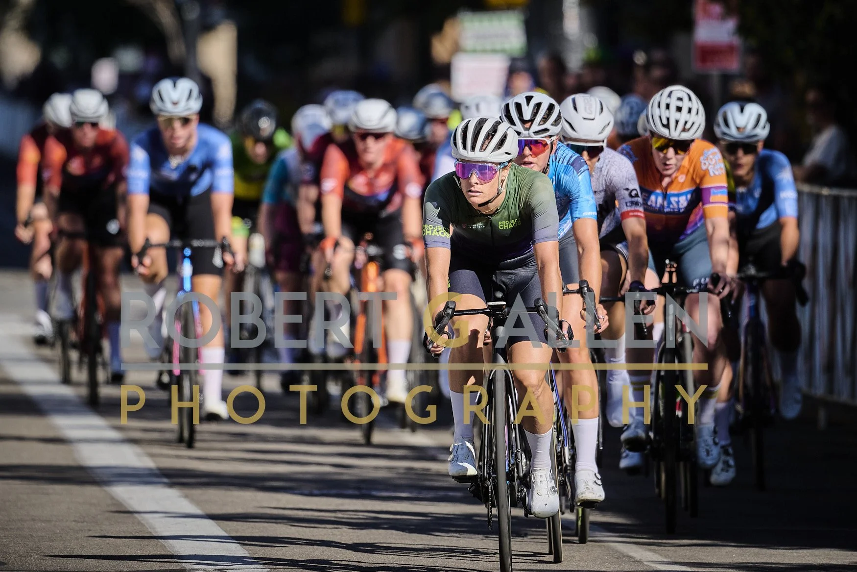 Cyclists participating in a race ride closely together on a city street, wearing helmets and colorful athletic gear.
