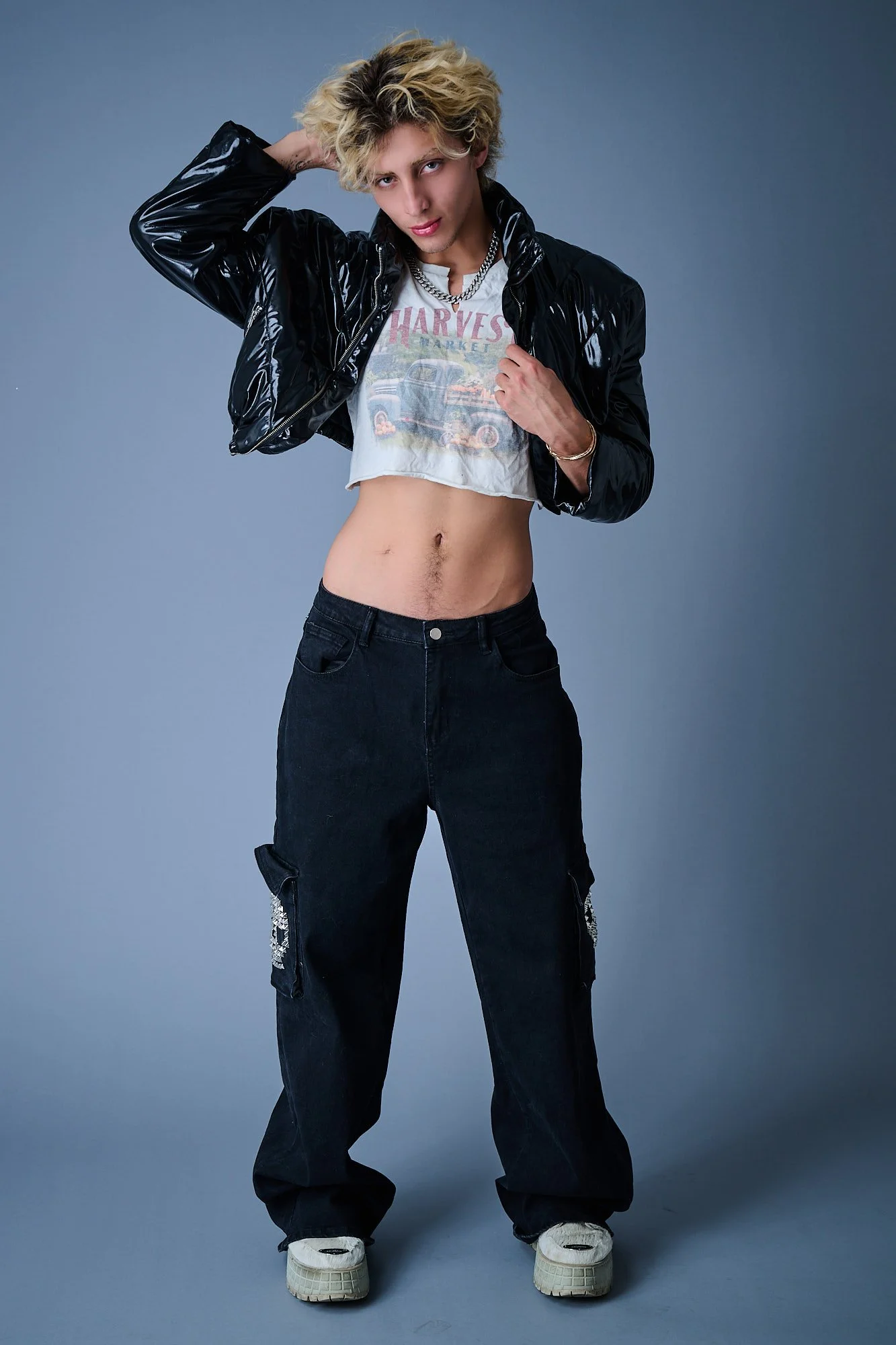 Young person with curly blonde hair wearing a black vinyl jacket, a cropped graphic T-shirt, baggy black pants, and white sneakers, standing against a gray background, posing with one hand behind their head.