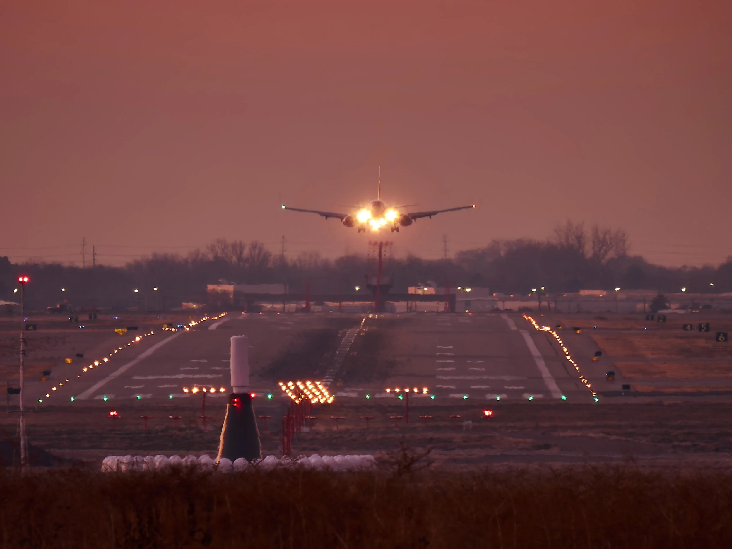 An airplane is taking off or landing on a runway at dusk, with bright lights and a pinkish sky.