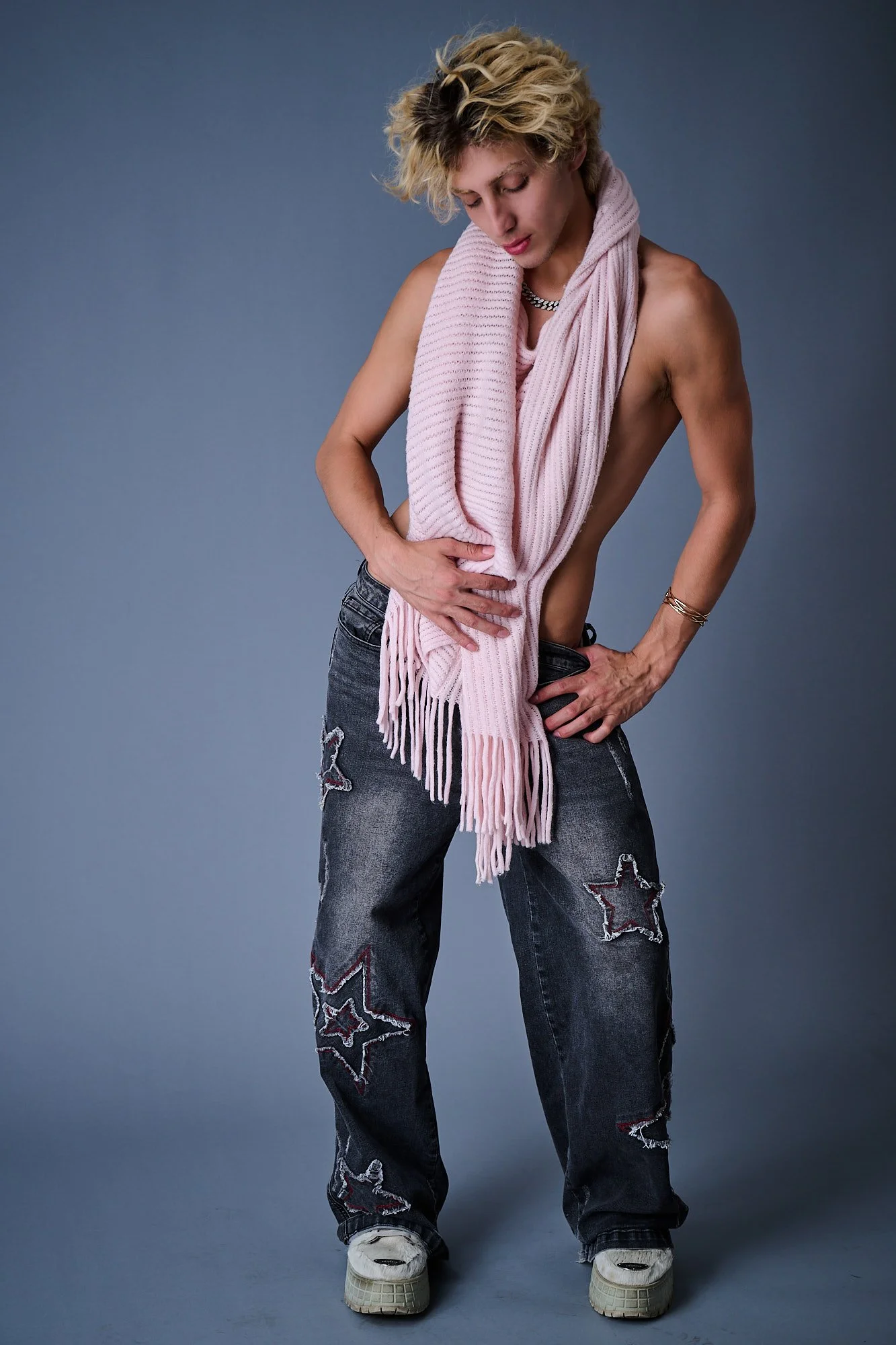 Person with blonde curly hair wearing ripped jeans, white sneakers, a pink scarf draped around shoulders, and a silver necklace, standing against a gray background.