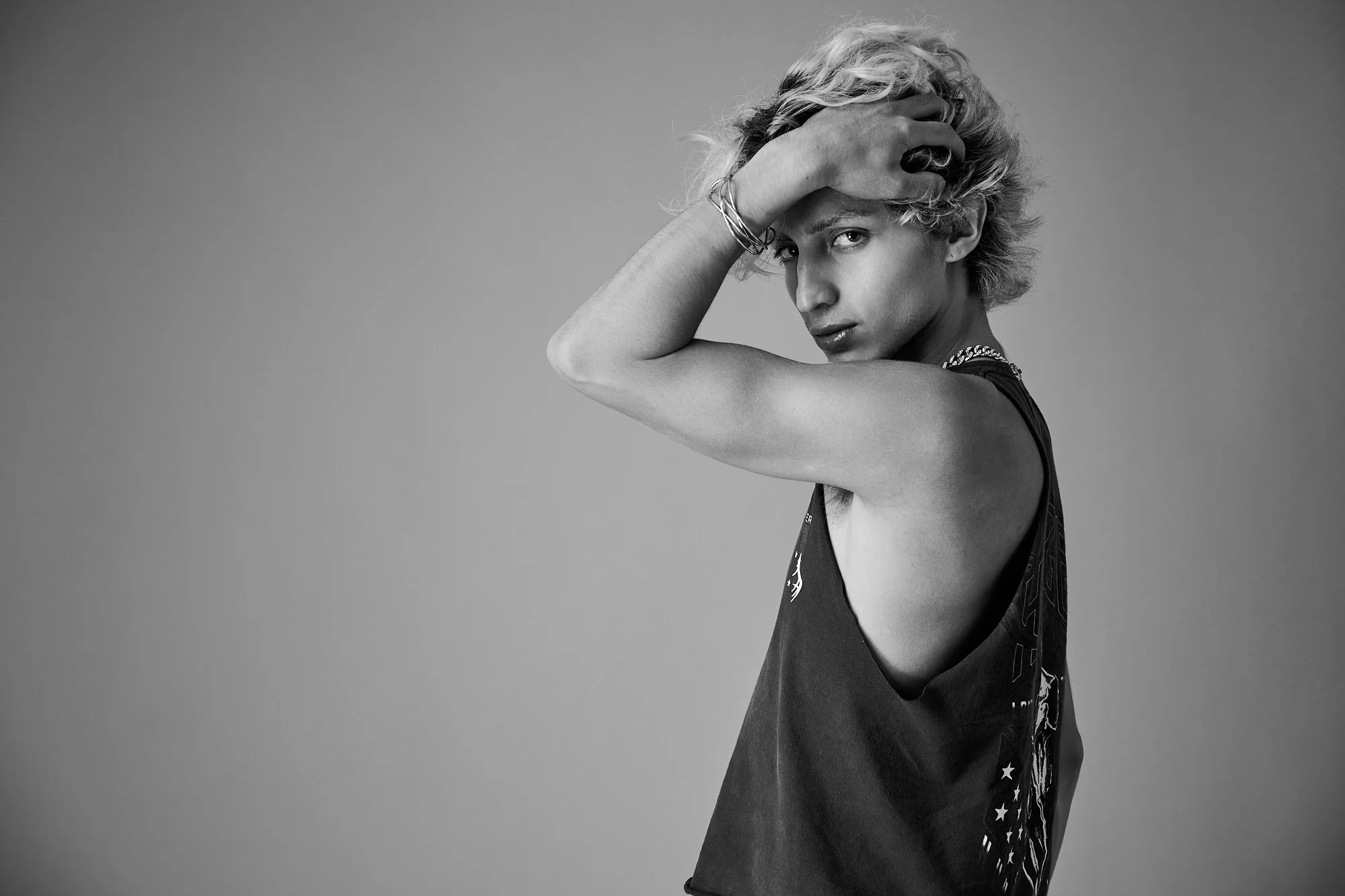 Black and white portrait of a young woman with short, curly hair, wearing a sleeveless shirt with stars and other patterns, holding her hand to her forehead, looking directly at the camera.