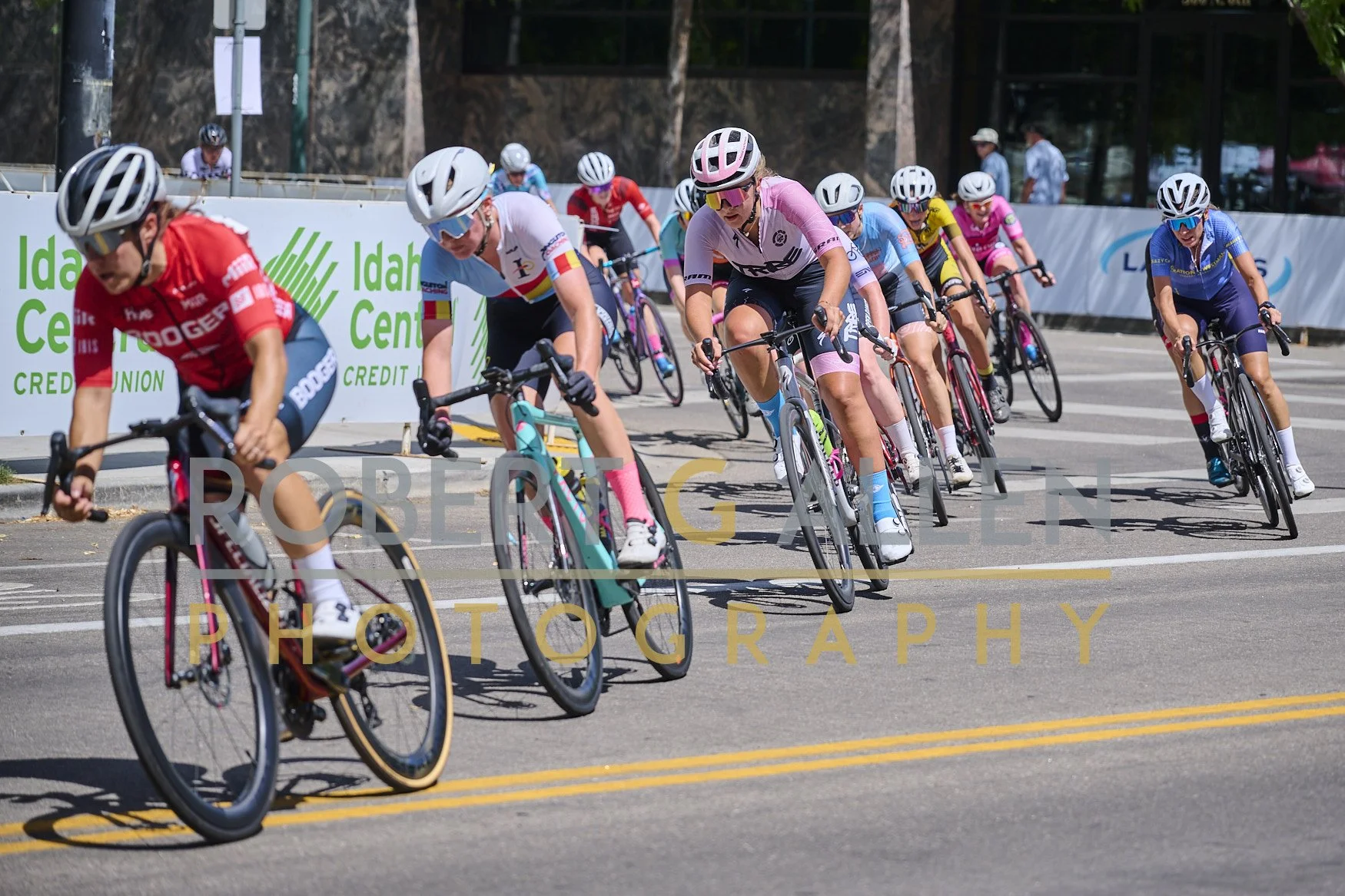 Group of female cyclists competing in a race on a city street, wearing colorful jerseys and helmets, with a green and white sign in the background.