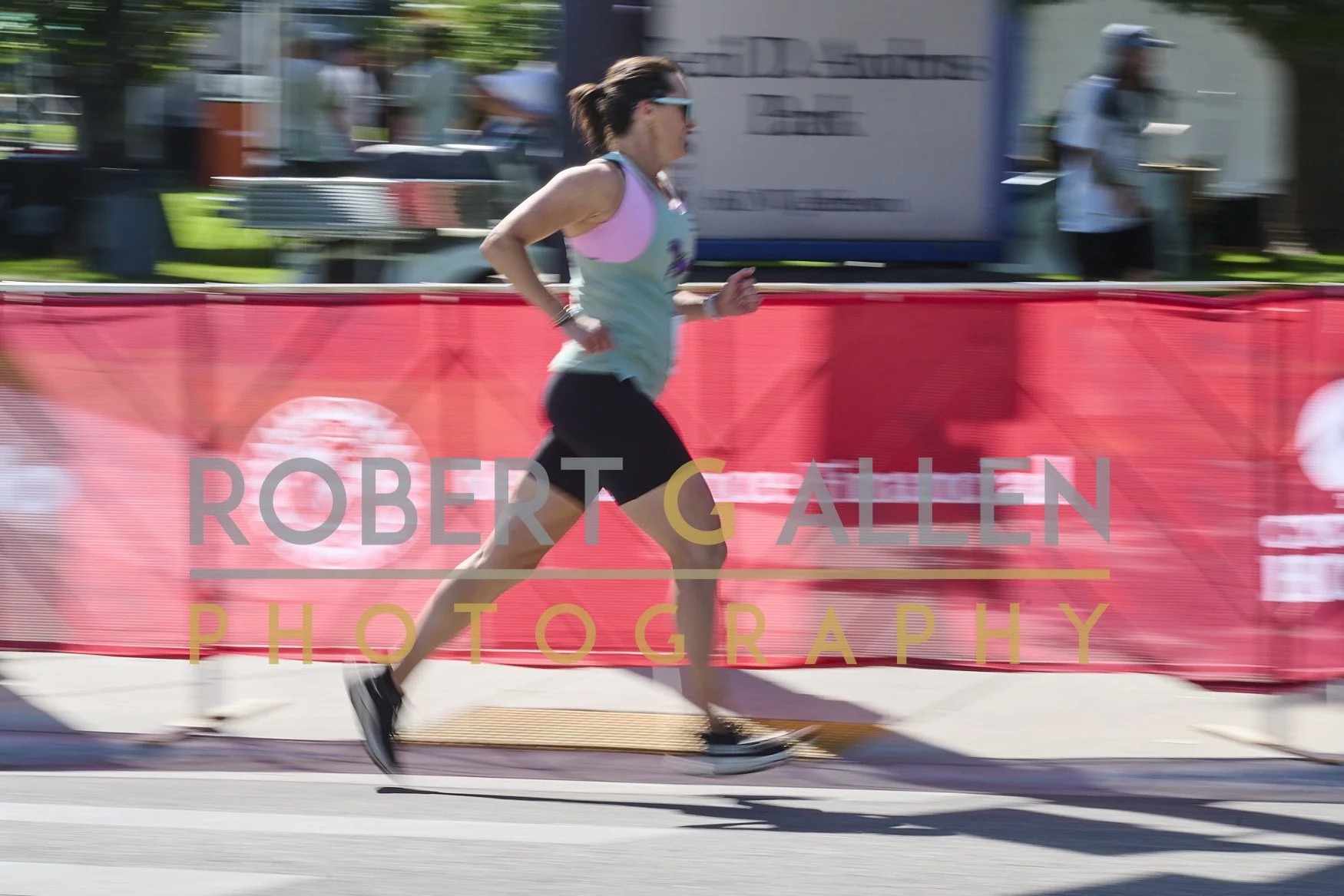 A woman running in a marathon race, wearing sunglasses, a sleeveless top, and black shorts, with a red safety barrier and an outdoor event setting in the background.