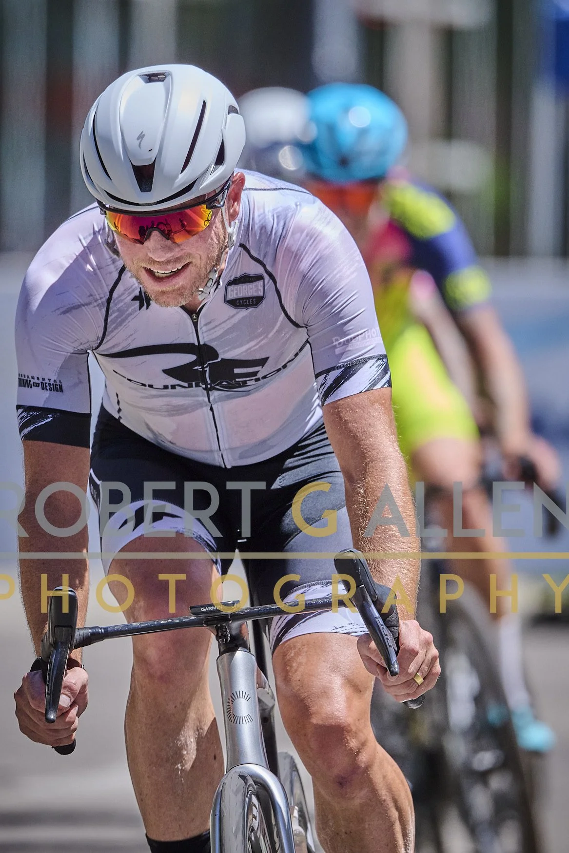 A man wearing a white cycling jersey, helmet, and sunglasses riding a bicycle, with a woman in colorful cycling attire and helmet riding behind him.