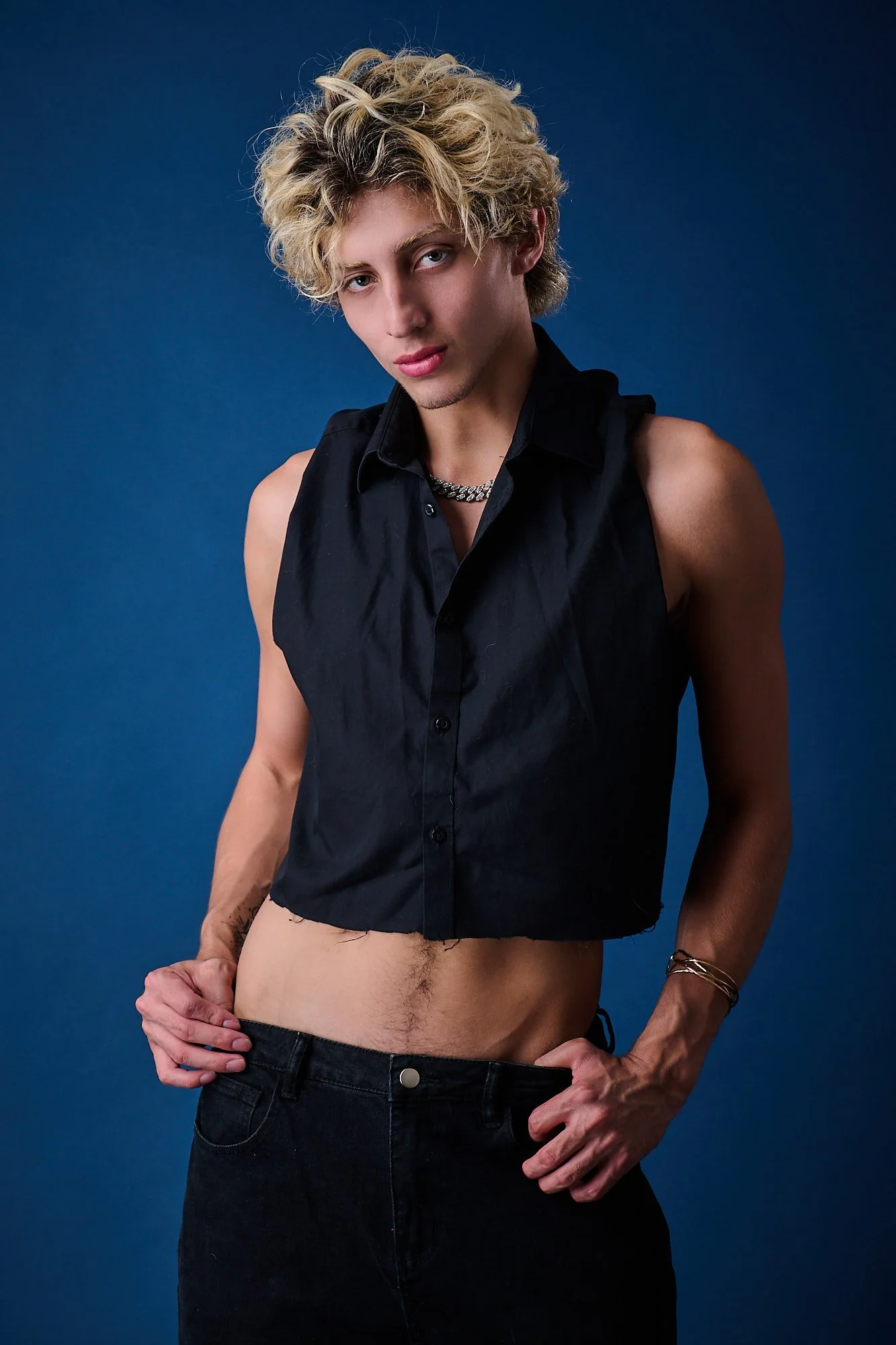 Young man with curly blonde hair, wearing a sleeveless black shirt, black pants, and necklaces, standing against a dark blue background.