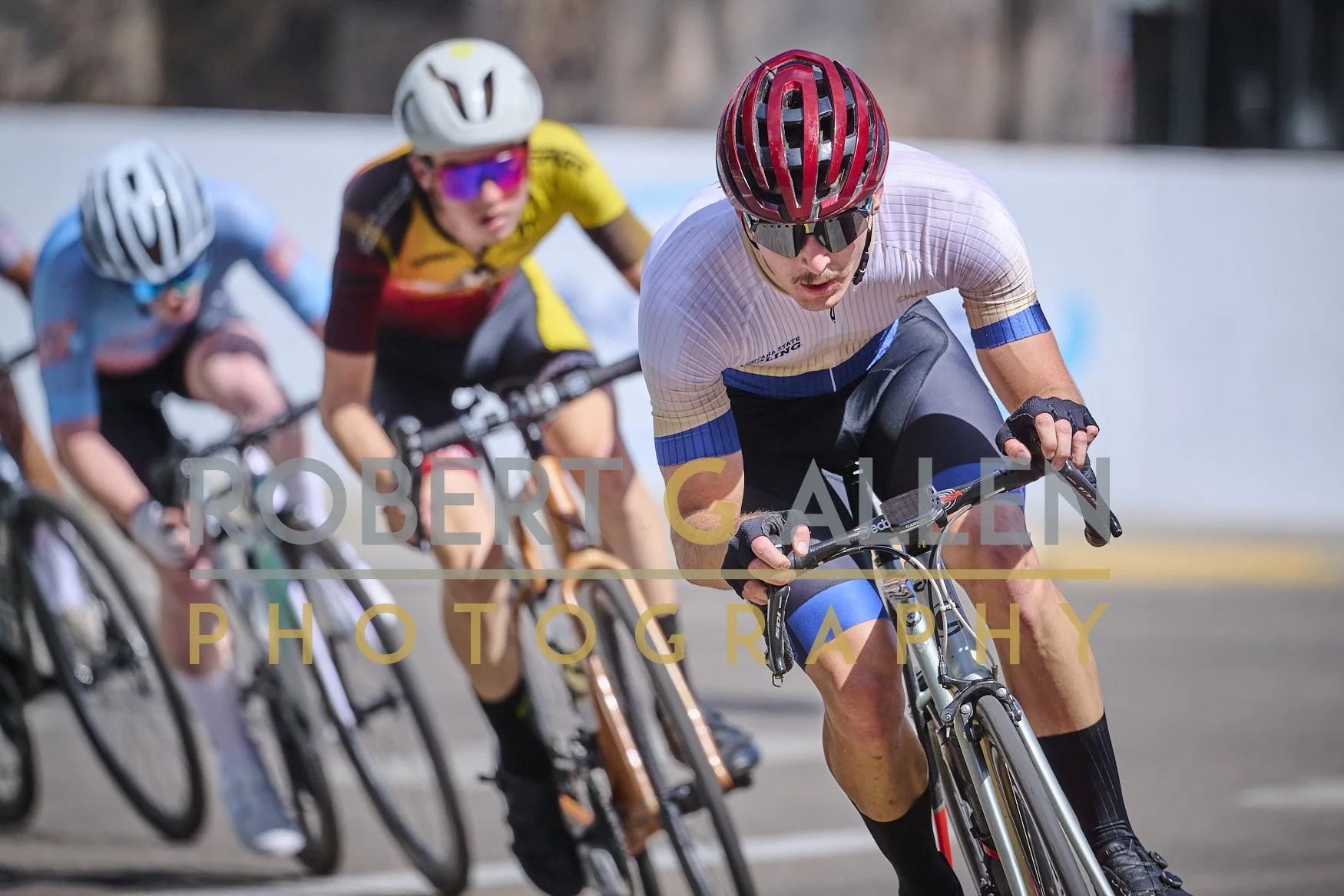 Cyclists racing on a road, wearing helmets and sports gear, with the focus on the lead rider in a white jersey and red helmet.