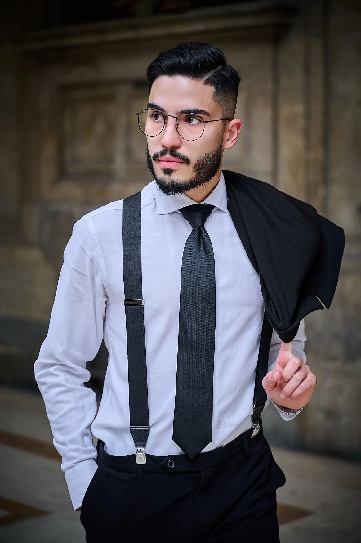 A well-dressed man with dark hair and beard, wearing glasses, a white shirt, black tie, suspenders, and carrying a black jacket over his shoulder, standing in front of a textured wooden background.