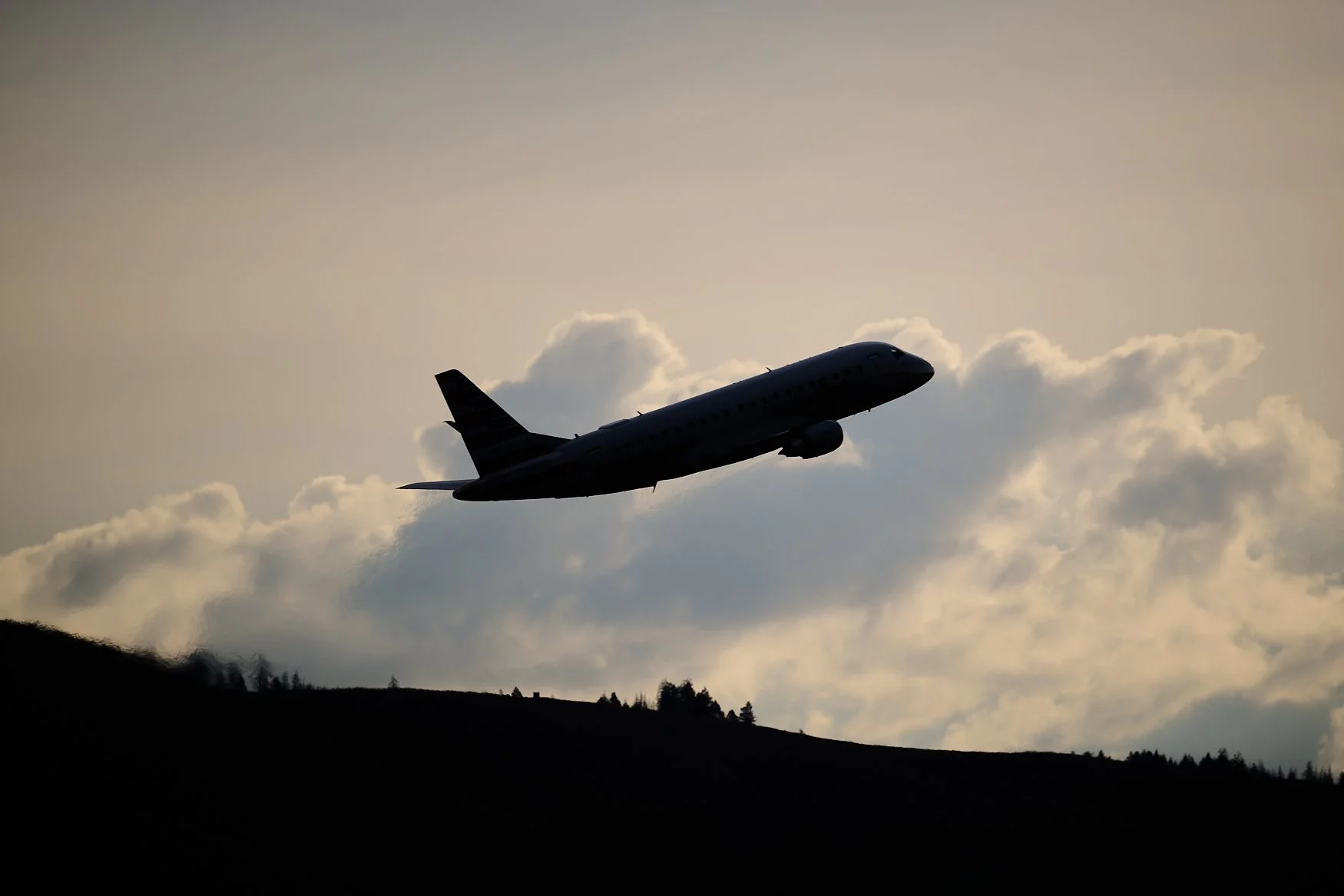 A silhouette of a commercial airplane flying in the sky with clouds and hills below.
