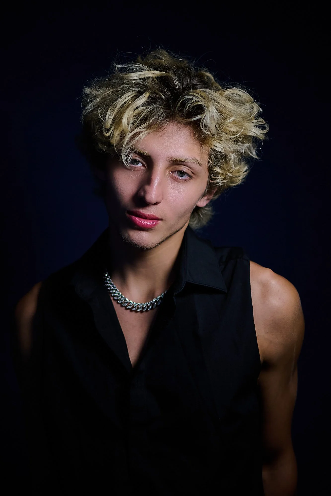 Portrait of a young person with curly blond hair, wearing a black sleeveless shirt, a silver chain necklace, against a dark background.