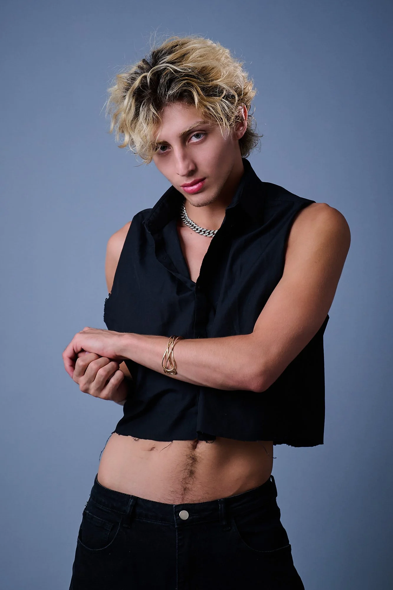 A young man with curly blonde hair and blue eyes wearing a black sleeveless shirt, a diamond necklace, and gold bracelets, posing against a plain blue background.