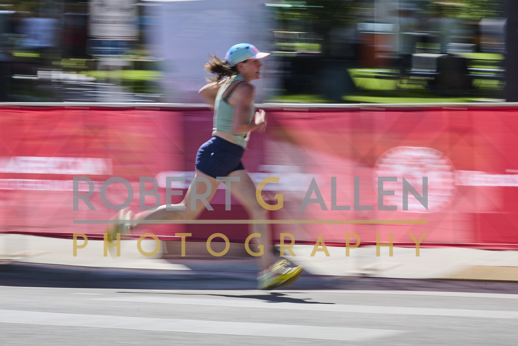 A woman running outdoors during a race, wearing a light blue tank top, dark shorts, a colorful cap, and running shoes, with a pink barrier and green park background.