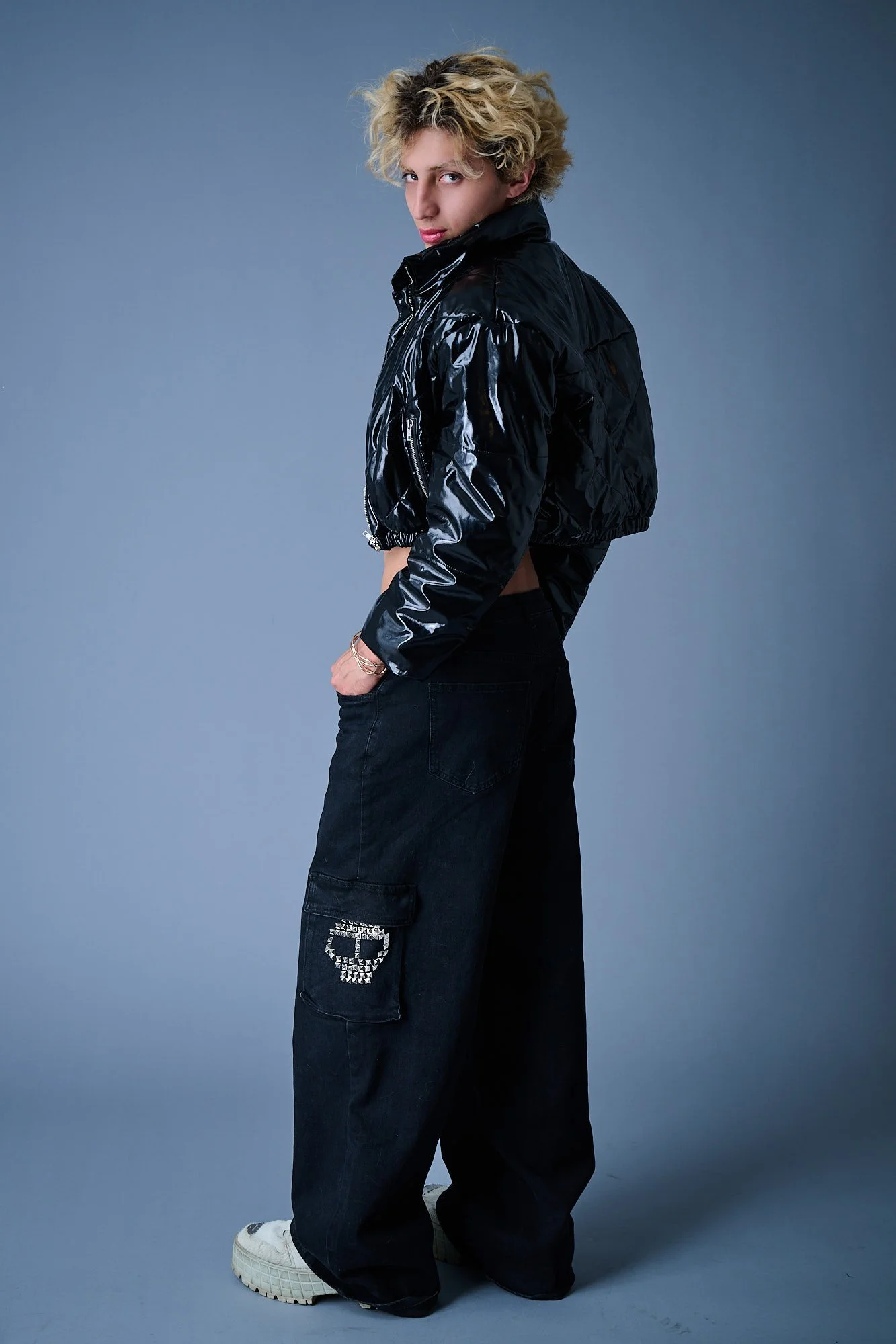 Person with blonde curly hair wearing a shiny black jacket, baggy blue jeans with a skull design on the pocket, and white sneakers, standing against a gray background.