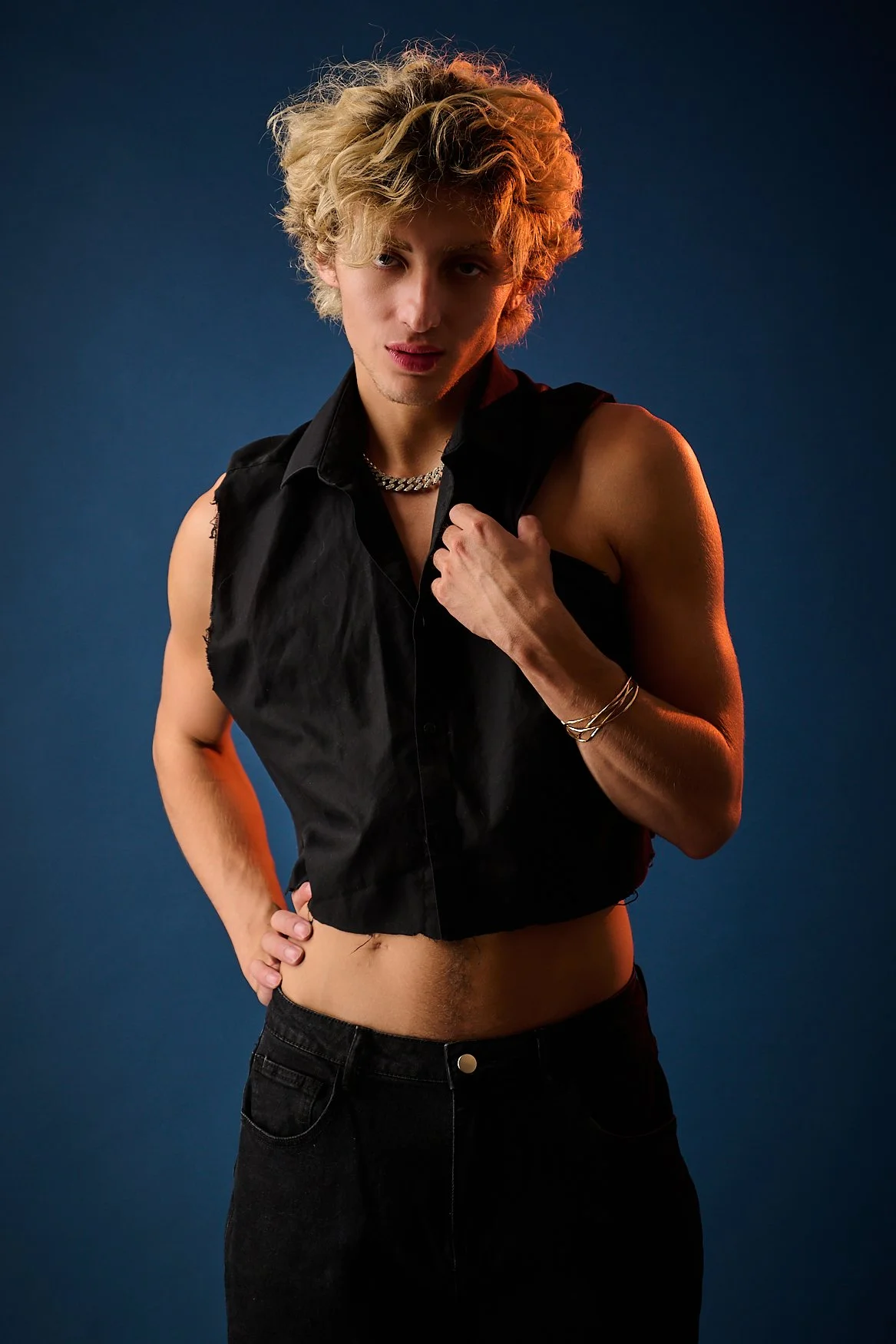 Person with curly blonde hair posing against a dark background, wearing a sleeveless black shirt, black pants, multiple jewelry pieces, and showing their midriff.