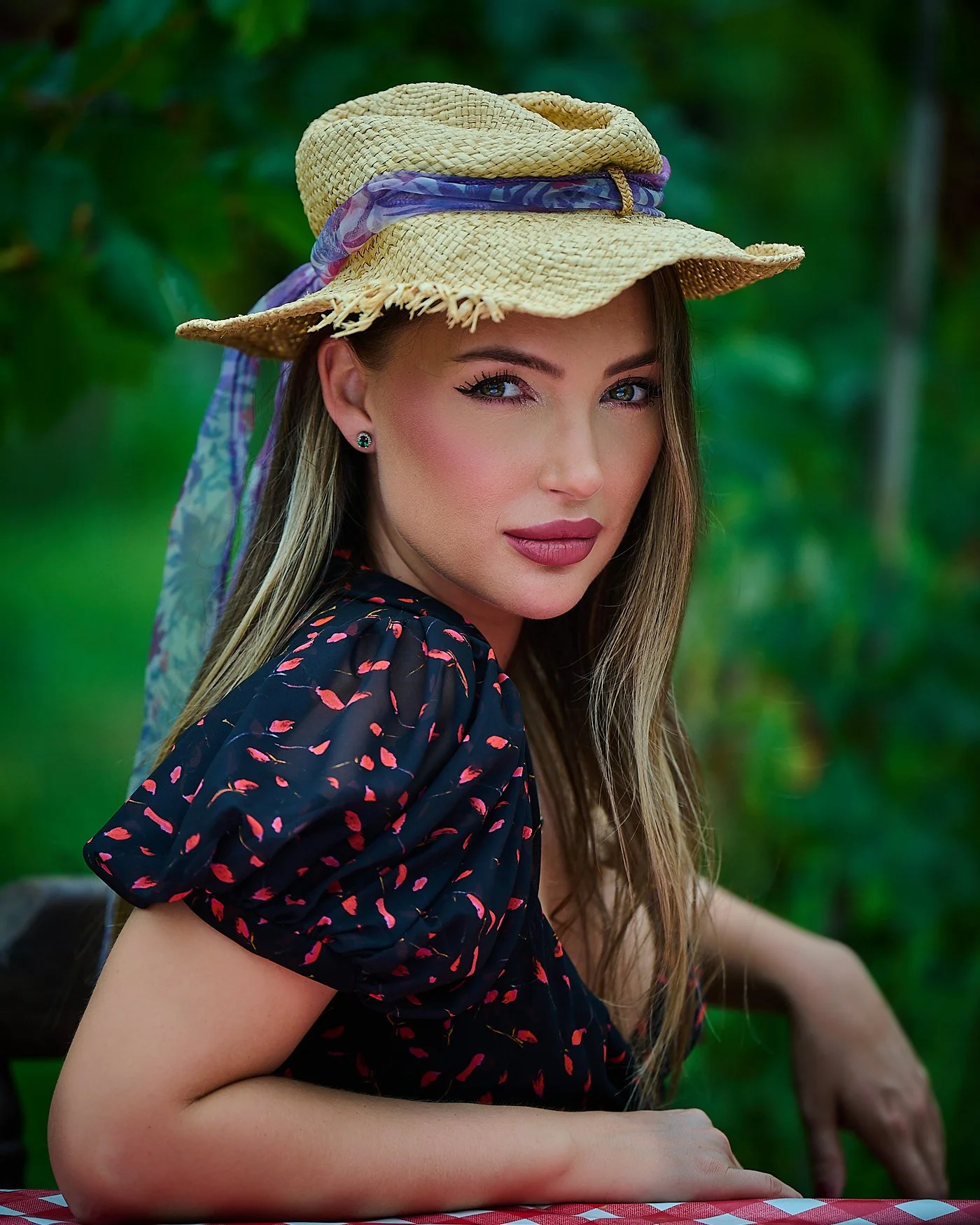 A woman wearing a straw hat with a colorful scarf band, sitting outdoors with a blurred green background. She has blonde hair, hoop earrings, and wears a dark dress with pink and purple patterns.