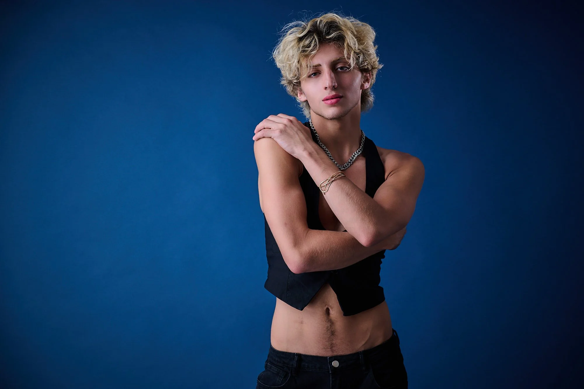Young person with curly blonde hair posing against a solid blue background, wearing a black sleeveless top, gold jewelry, and black pants.