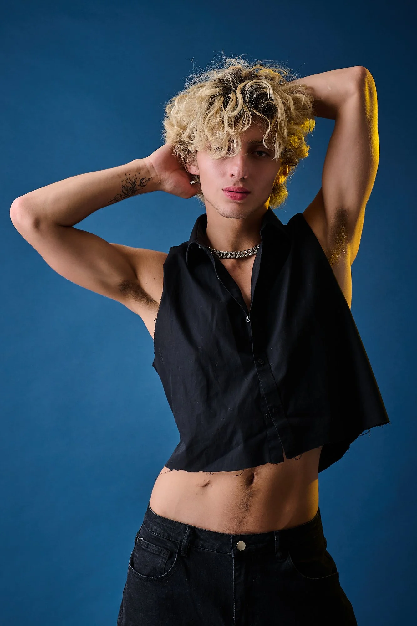 Young man with curly blonde hair posing against a solid blue background, wearing a sleeveless black shirt, black jeans, a chain necklace, and a tattoo on his left arm.