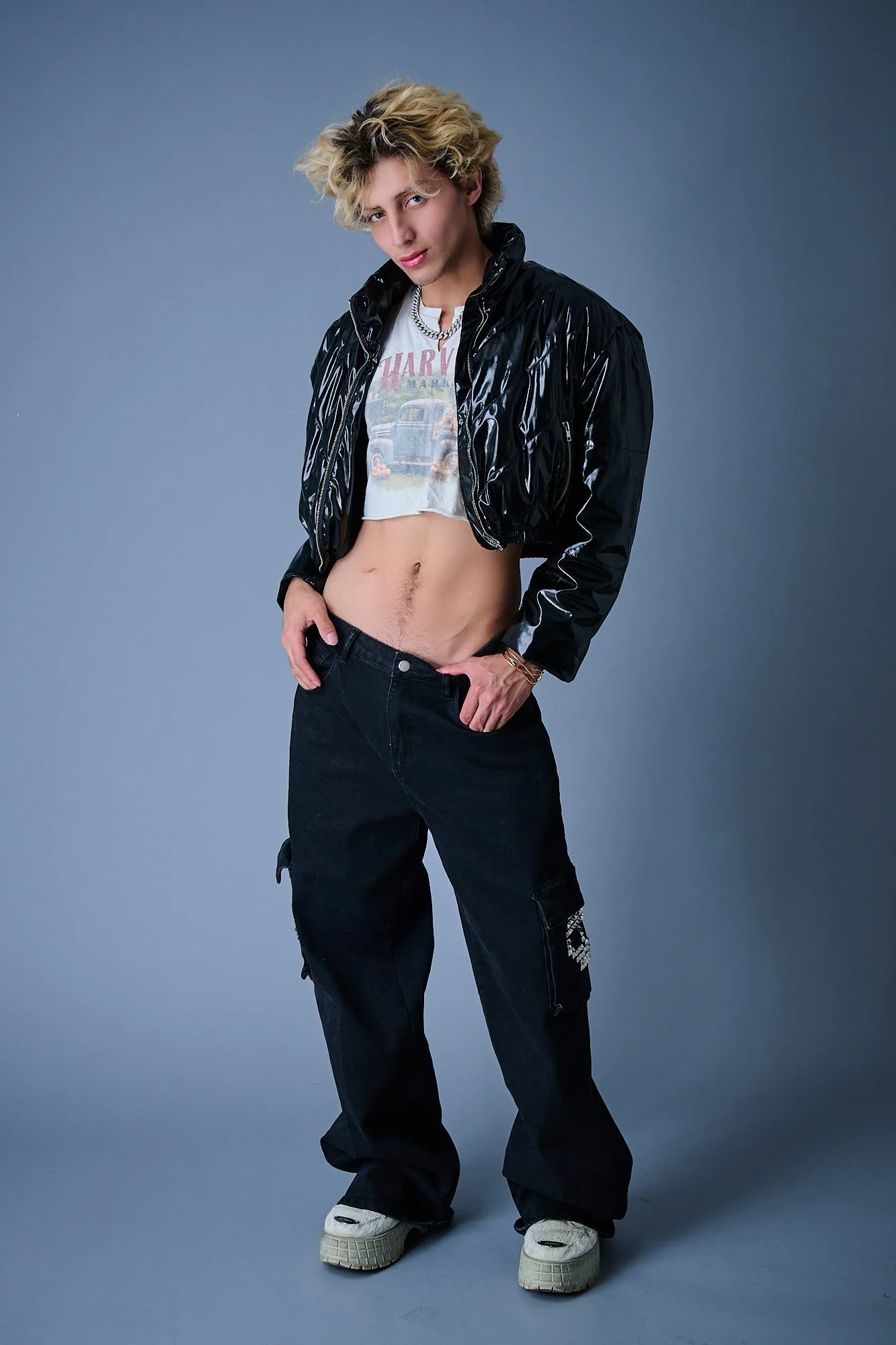 A young person with wavy blonde hair wearing a black shiny jacket, a vintage Harley-Davidson T-shirt, black cargo pants, and sneakers, posing against a gray background.