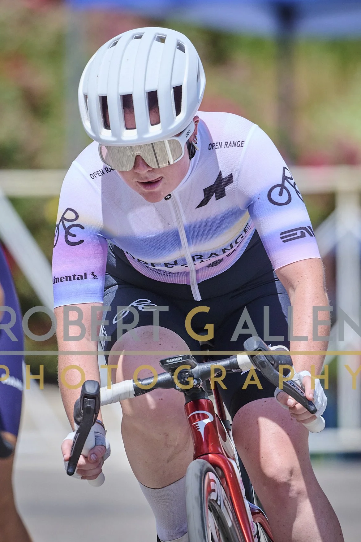 A cyclist wearing a white helmet, sunglasses, and a white cycling jersey is riding a red bicycle during a race, with a showing focused expression.