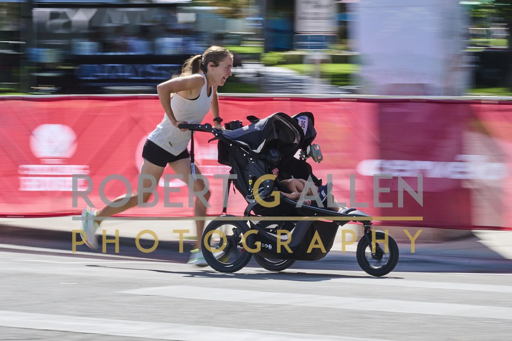 A woman participating in a race pushes a jogging stroller with a child inside, running on a city street with a red barrier and blurred background.