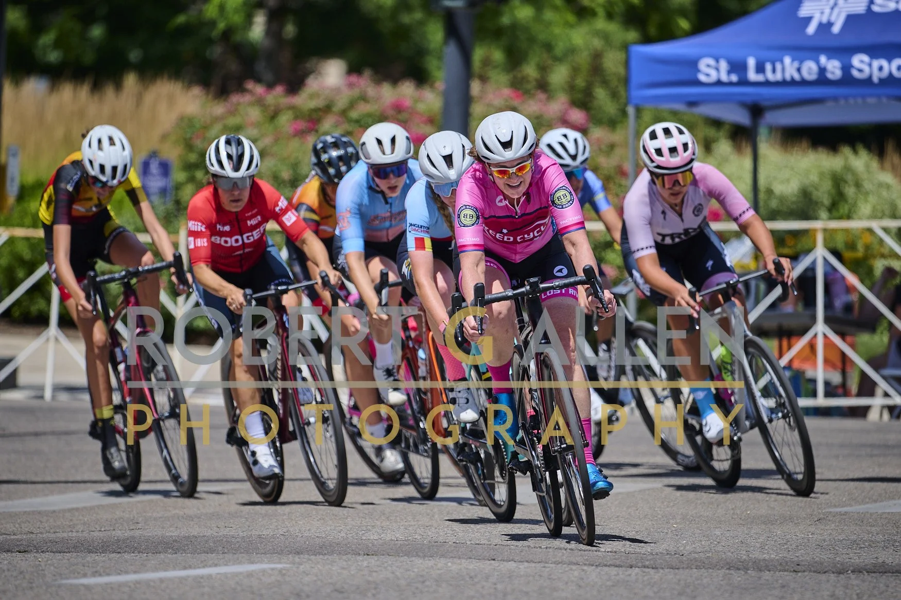 Women cyclists in a race wearing helmets and colorful jerseys, riding on a paved road with barriers, trees, and a tent in the background.