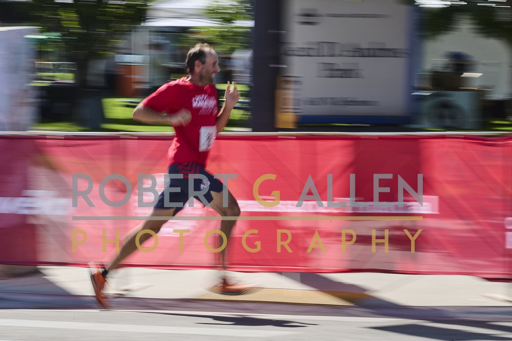 A man running in a race, wearing a red shirt and black shorts, with a race number pinned to his shirt. He is outside, with green trees and buildings in the background, and a red barrier with the text "Robert G Allen Photography" in front of him.