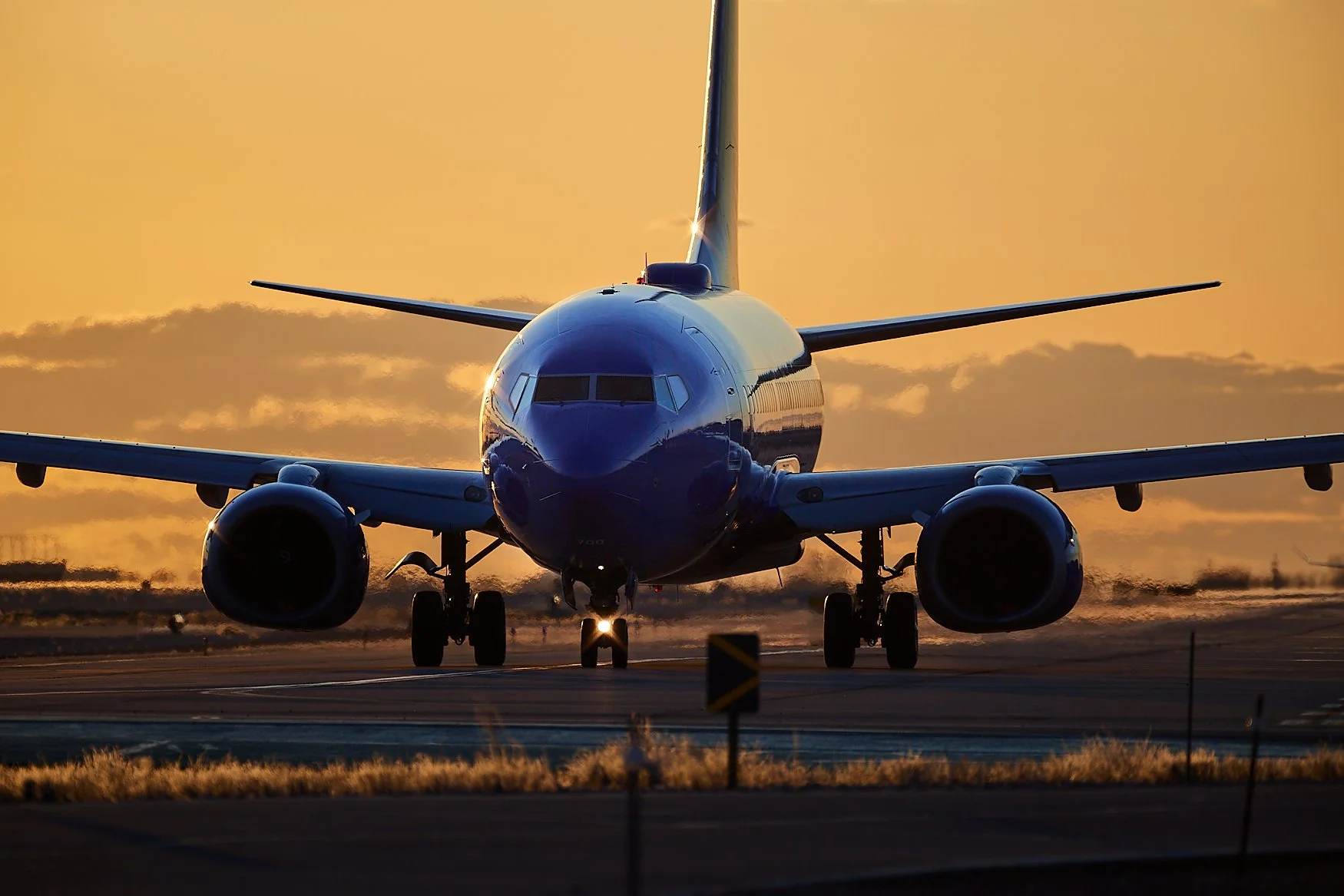 A large commercial airplane on the runway during sunset, with the sky painted in warm orange tones and the plane's front facing the camera.
