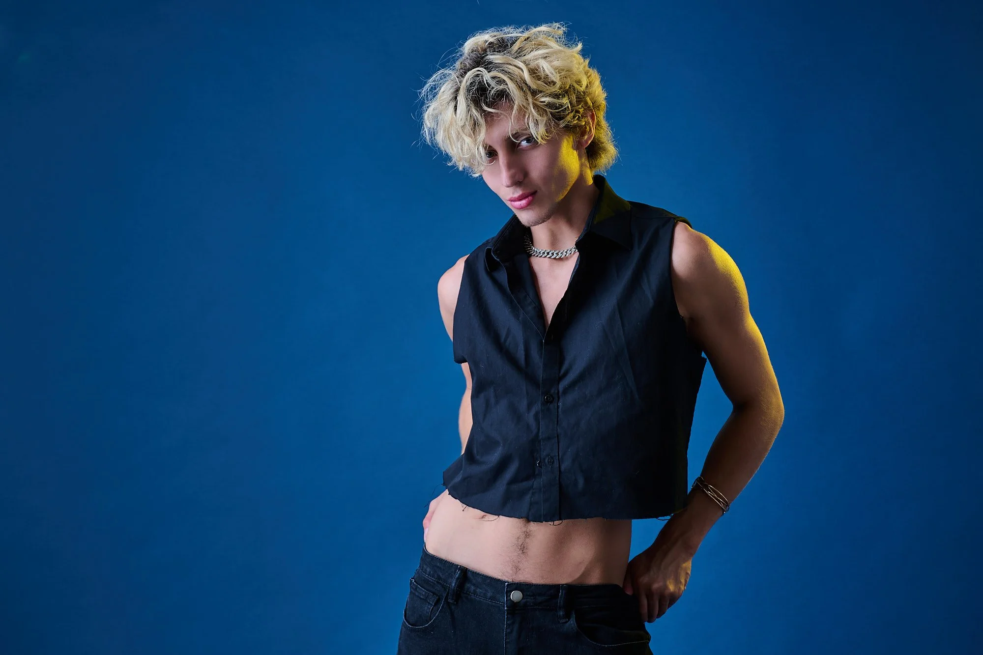 A young man with curly blond hair wearing a sleeveless black shirt, chain necklace, and jeans, standing against a blue background.