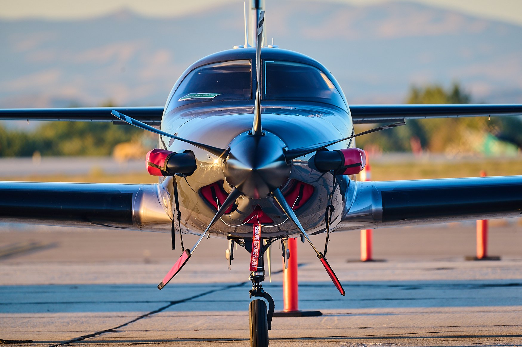 Front view of a small aircraft on the runway during daytime, with orange cones around it and a warning strap on the nose gear.