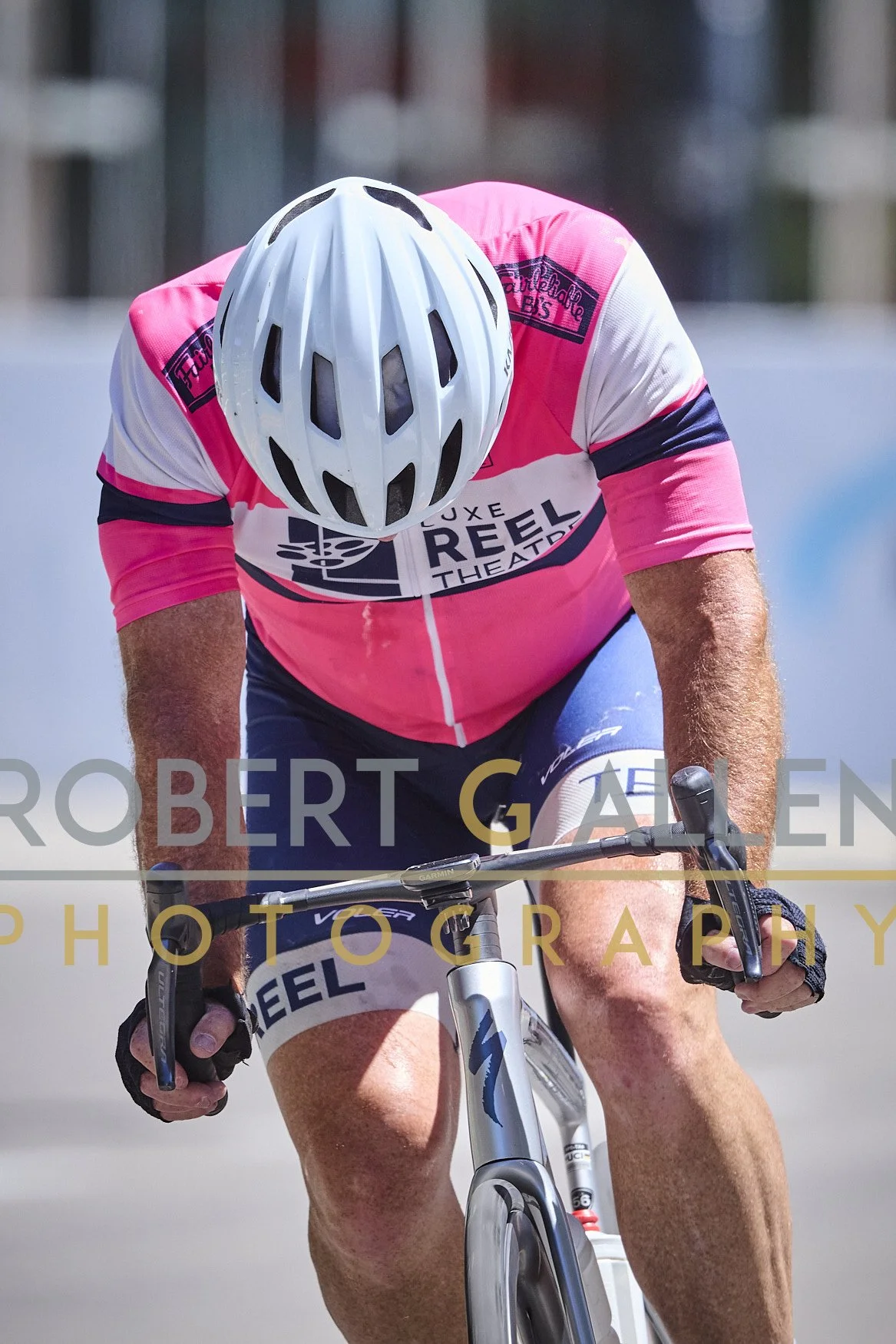 A male cyclist wearing a pink and white jersey with a white helmet, focused and leaning forward on a mountain bike during a race or ride.