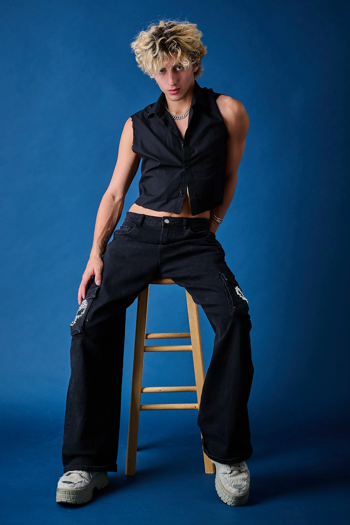 Person with curly blond hair wearing a black sleeveless shirt, dark jeans, and white sneakers, sitting on a wooden stool against a blue background.
