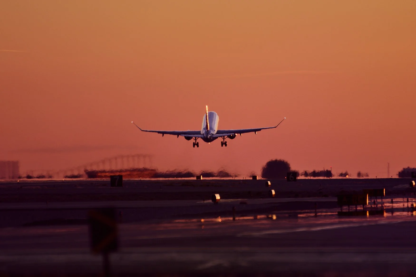 An airplane landing during sunset at an airport runway, with a pink-orange sky and orange runway lights.