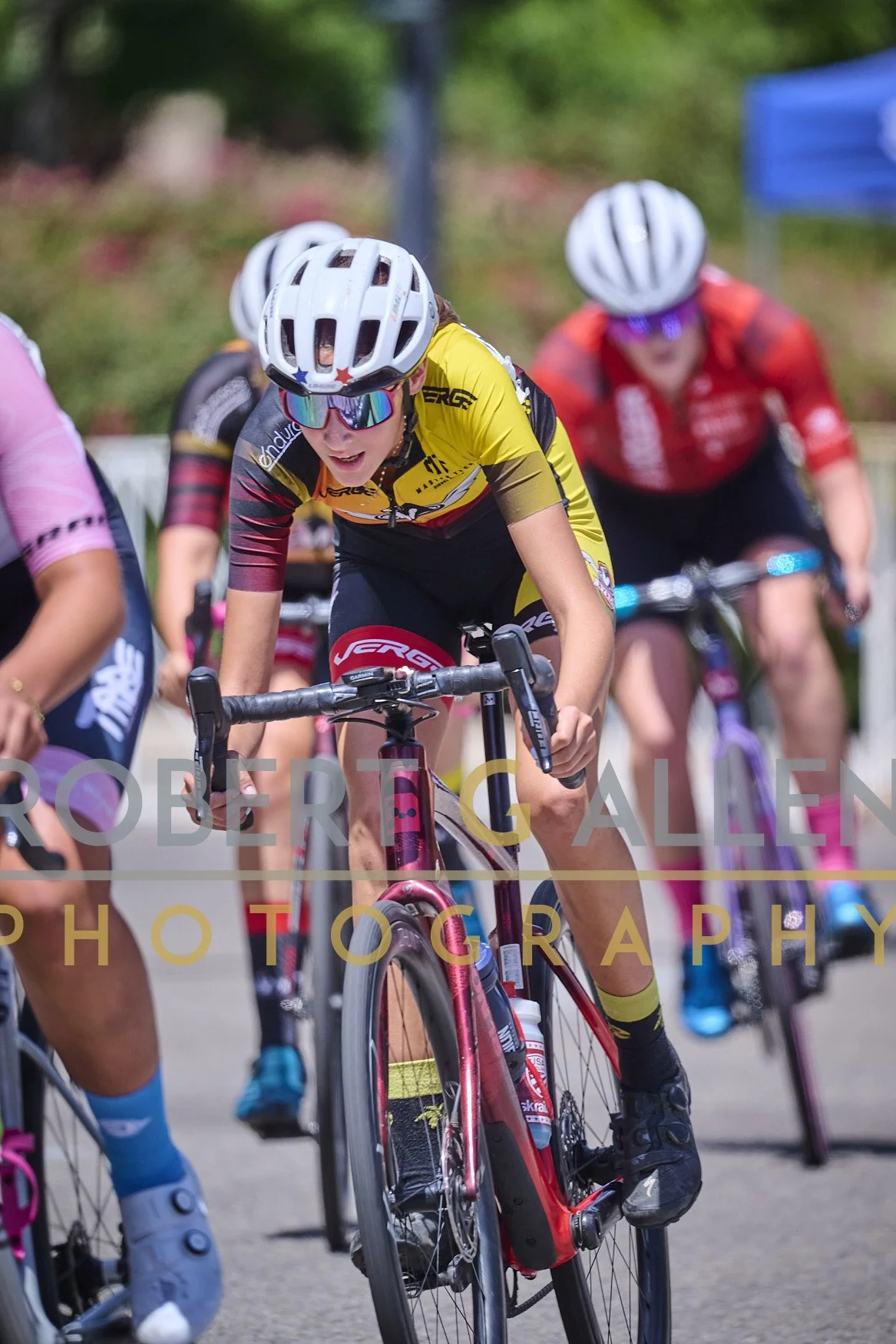 Cyclists racing, with one woman in yellow and black jersey wearing a white helmet and sunglasses, riding a pink mountain bike.