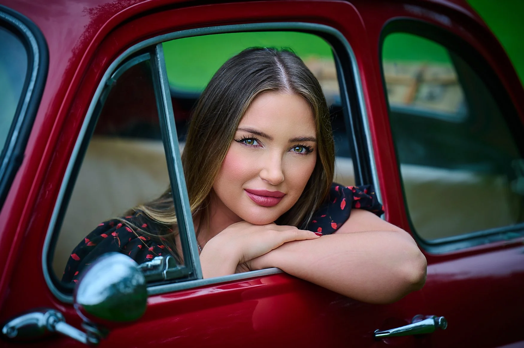 A woman with long brown hair and blue eyes leaning on the window of a vintage red car, resting her chin on her arms, and smiling gently.