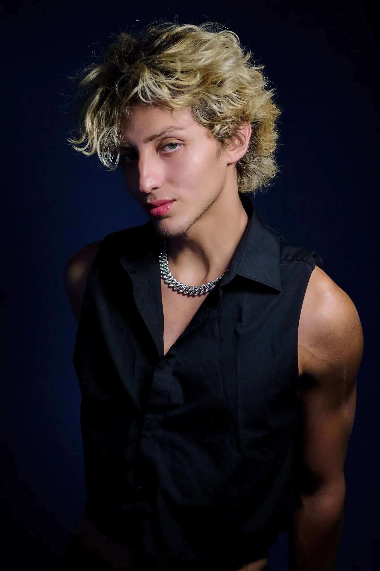 Young man with curly blond hair, wearing a black sleeveless shirt and a silver chain necklace, posing against a dark background.