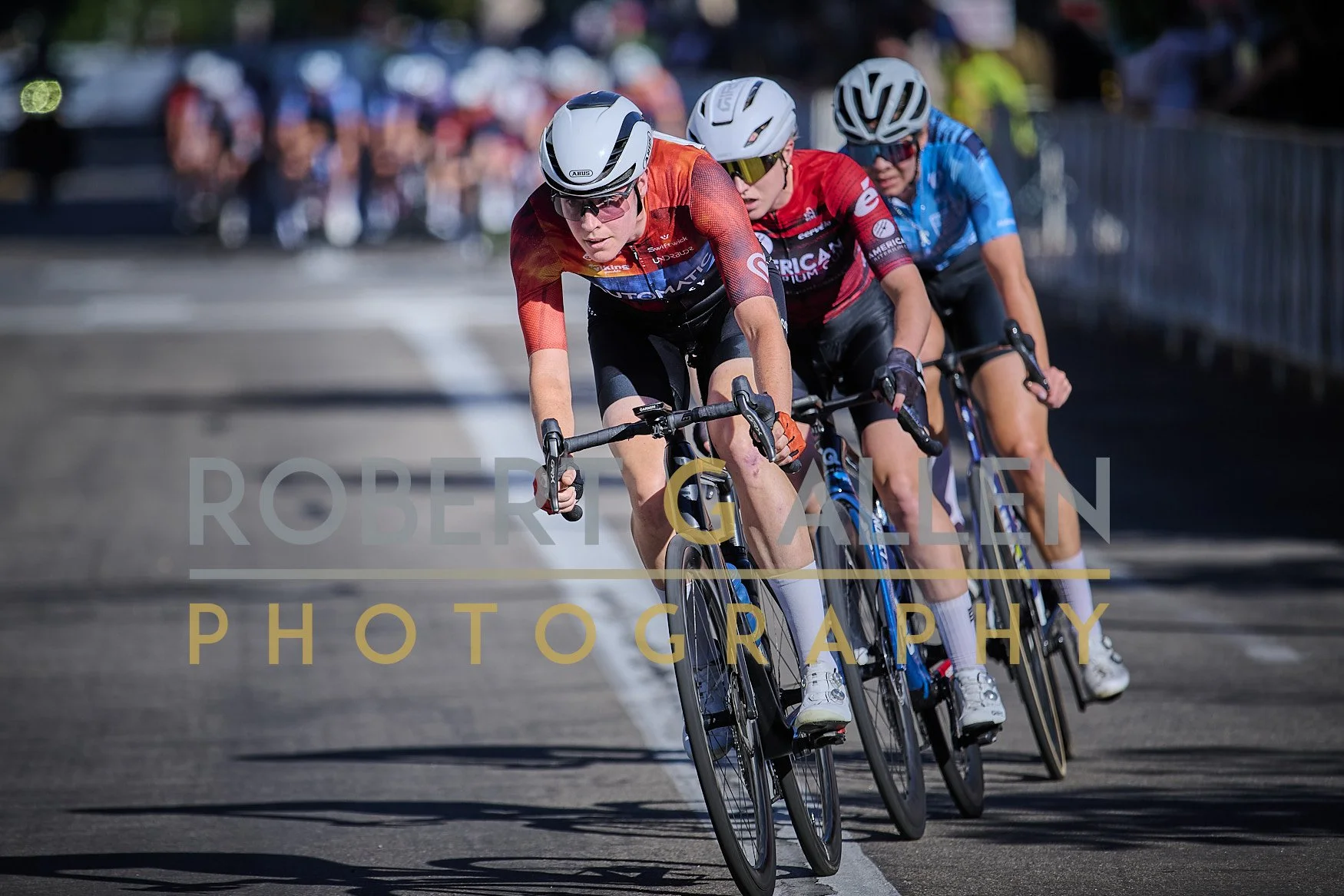 Cyclists racing in a bike race, wearing helmets and athletic gear, leaning into a turn on a paved road with spectators in the background.