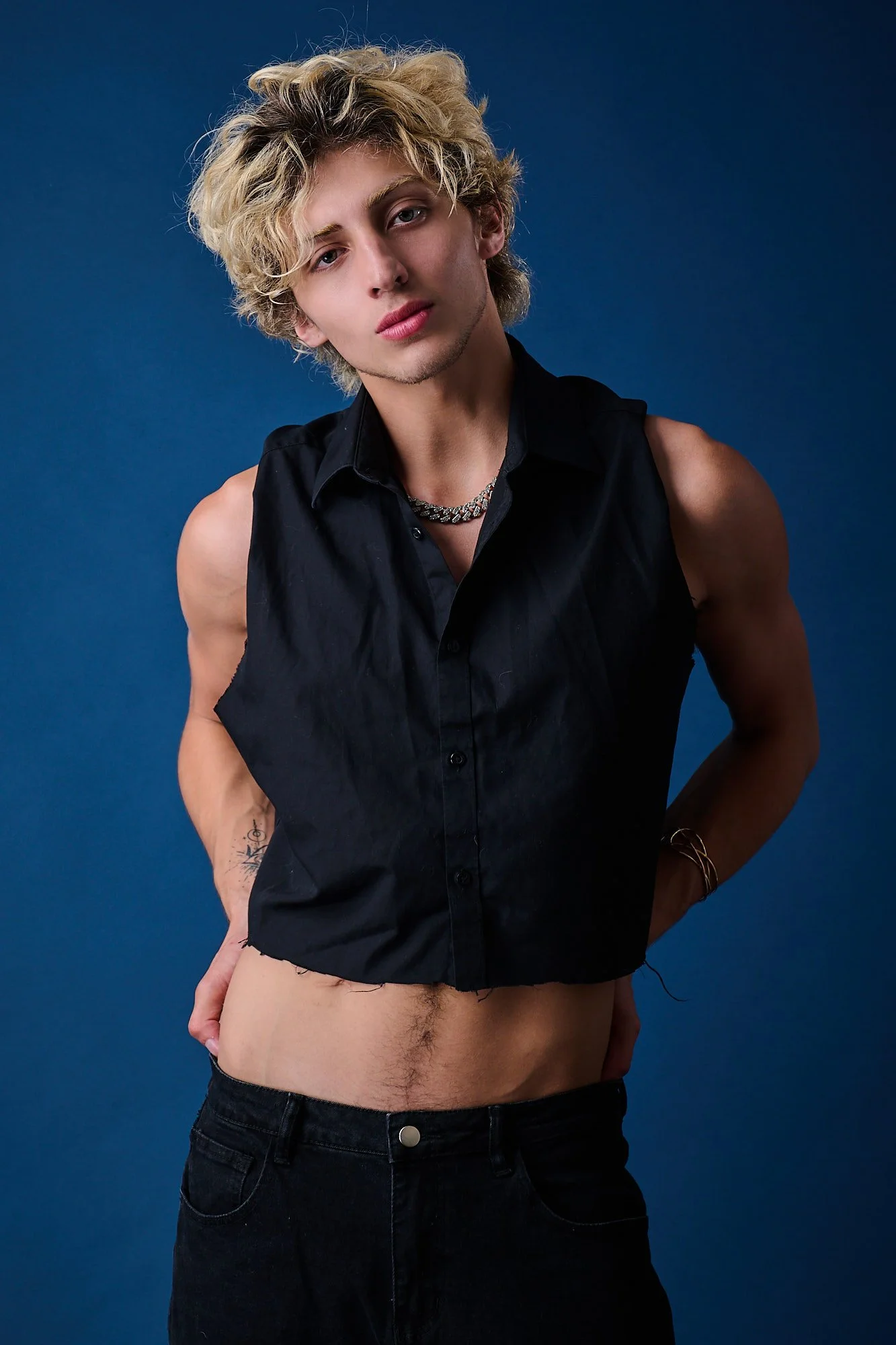 A young man with blonde, curly hair and light skin, wearing a sleeveless black shirt, a chain necklace, bracelets, and dark jeans, standing against a solid blue background.
