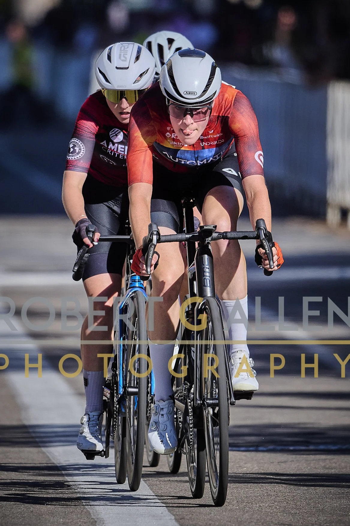 Two cyclists in a race, wearing helmets, sunglasses, and athletic clothing, riding on a paved road with a person in the background.