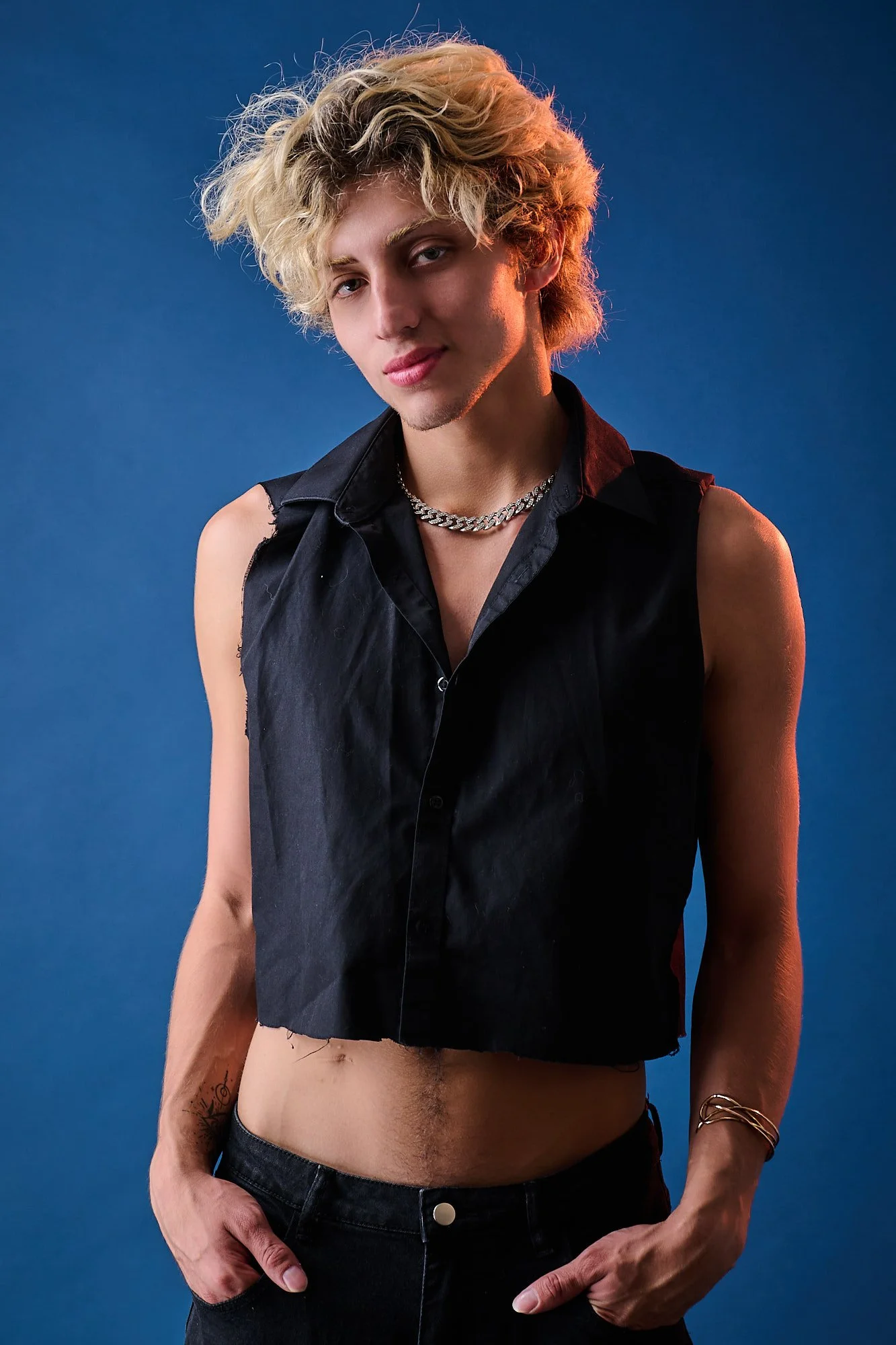 A young man with bleach blonde, curly hair, wearing a black sleeveless vest, a silver chain necklace, dark jeans, and a gold bracelet, standing against a blue background with a confident expression.