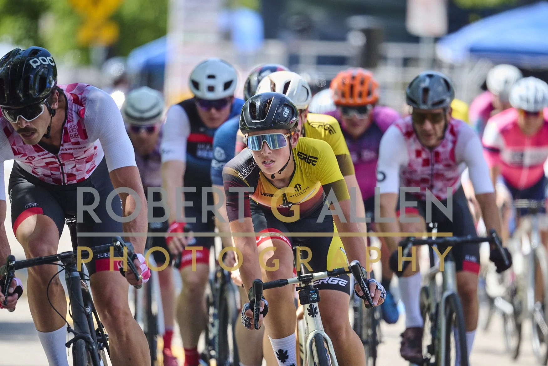 Group of cyclists participating in a race, wearing helmets and sports gear, with a focus on a female cyclist in a yellow and black jersey at the front.